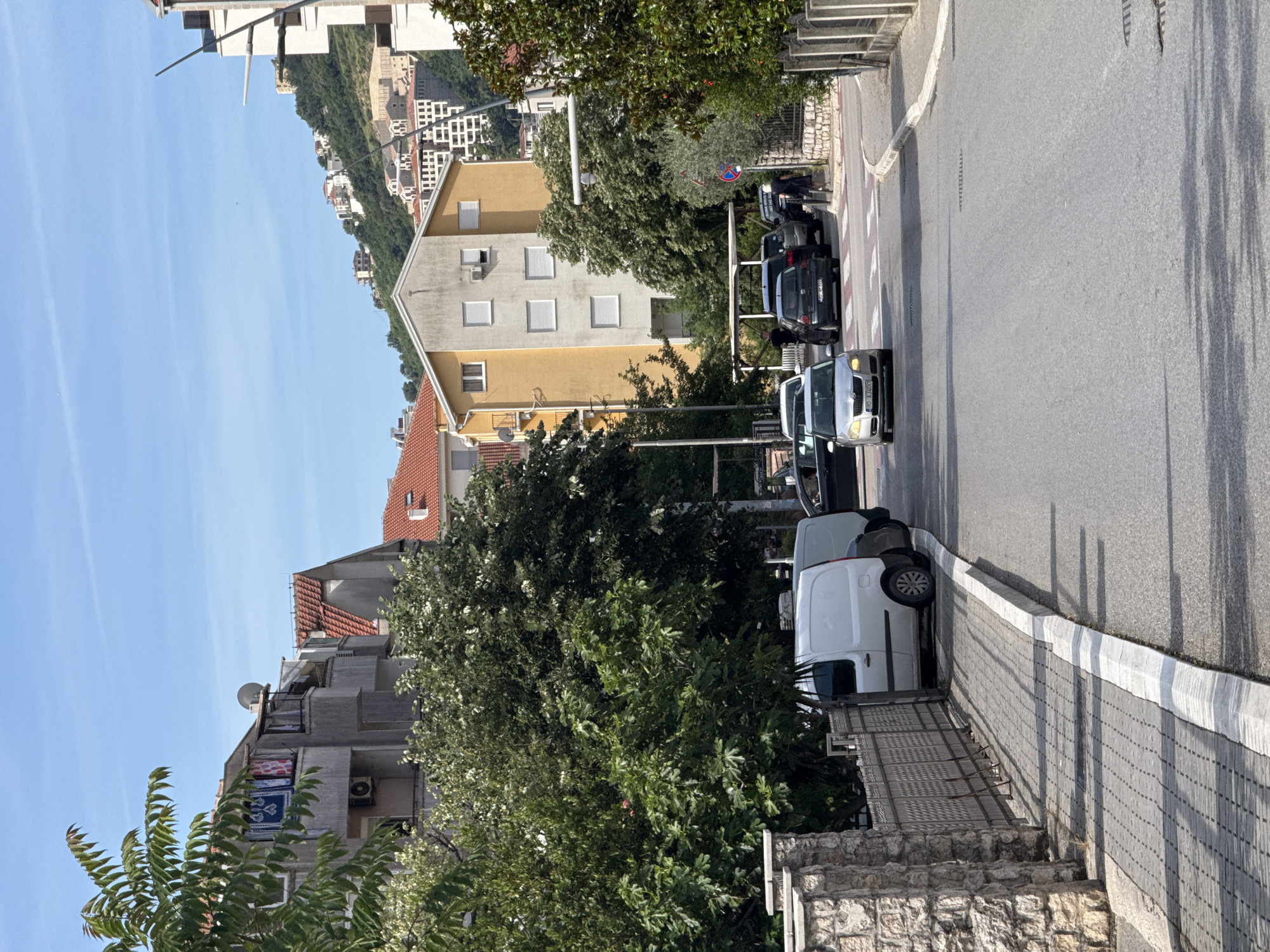 This media file captures a sunny street scene in what appears to be a residential area of Budva, Montenegro, consistent with the provided location context. The image is taken from a slightly elevated perspective, looking down a gently curving asphalt road.
**Subjects:**
The scene features several multi-story buildings. On the left, a light yellow/beige building with a grey roof and both open and shuttered windows is prominent. Further left and behind, a grey apartment building with multiple balconies, some displaying hanging rugs or laundry, is visible. In the distance, on a green hillside, numerous white or light-colored buildings are scattered.
A line of cars is parked along the right side of the road, including a white delivery van, a silver car (with license plate "BD 071-CO"), and a dark grey car (with a partial "BD 075" license plate visible). Another white van is further down the street.
Abundant green trees and bushes are integrated throughout the scene, some with white or pinkish blossoms, partially obscuring buildings and casting shadows.
The ground features a paved sidewalk with a distinctive white stripe along the curb, and stone walls and steps in the immediate foreground.
**Activity or Event:**
The image depicts a typical urban street during the day. Cars are parked, suggesting residents are at home or nearby. The hanging laundry implies everyday domestic life. There are no people visible, and no active events are taking place, creating a sense of calm.
**Notable Details:**
The time of day is bright, likely late morning or early afternoon, indicated by the clear blue sky and the well-defined shadows cast by trees and buildings on the road and sidewalk. The weather is clear and sunny. The architecture, with red-tiled roofs and hillside construction, is characteristic of the Mediterranean or Balkan region. A road sign indicating "no stopping" is visible behind one of the parked cars. The "BD" prefix on the license plates confirms the location as Budva.
**Visible Text:**
Visible text includes the license plate "BD 071-CO" on the silver car and partially "BD 075" on the dark grey car. A red circular no-stopping sign is also visible.