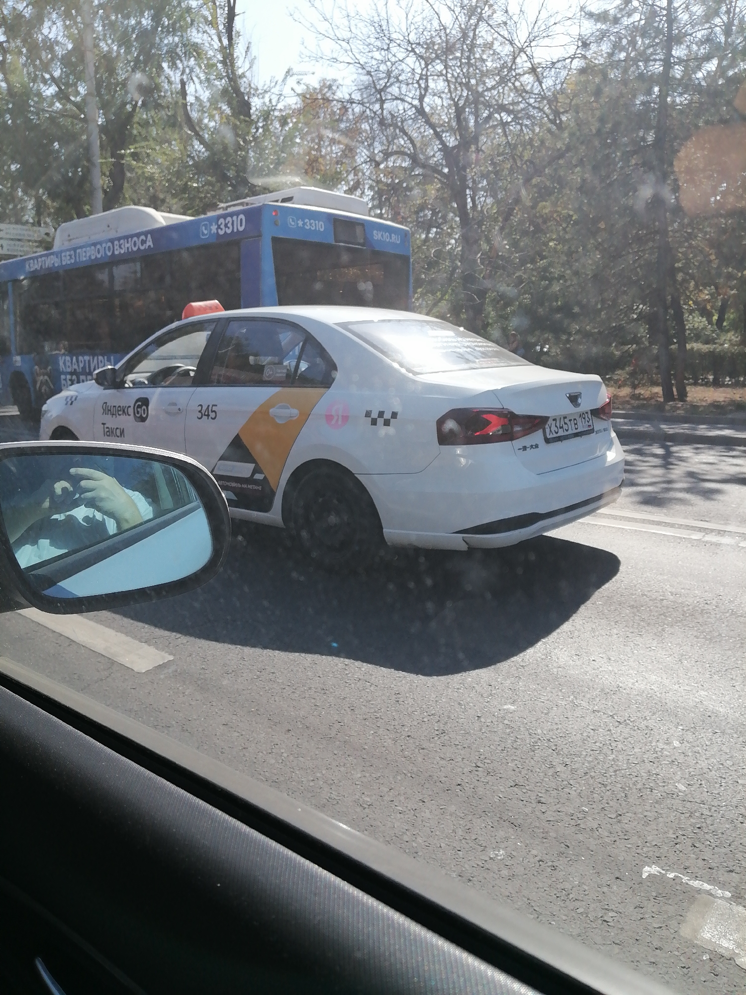 A white Yandex Go taxi is traveling on a paved road in Rostov-on-Don, Russia, possibly on a sunny day. The taxi has the Yandex Go logo and the number 345 on its side. In the background, a blue bus with the text "КВАРТИРЫ БЕЗ ПЕРВОГО ВЗНОСА" (Apartments without the first down payment) and "3310" is visible. The driver's hands are visible in the side mirror of the vehicle from which the photo is taken. The road surface shows a white line.
