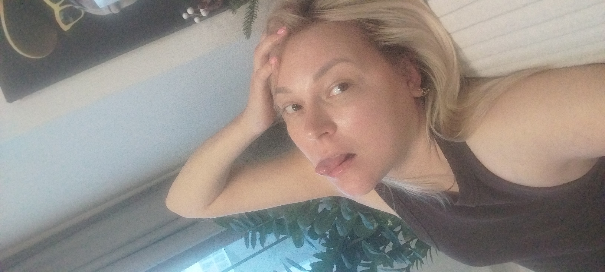 A woman with blonde hair, wearing a dark tank top, is lying down. Her left hand is resting on her forehead, and she is sticking her tongue out. The background includes a painting with glasses, some greenery, and what appears to be a window with plants outside. There is no indication of Izhevsk, Russia, or any other specific city.