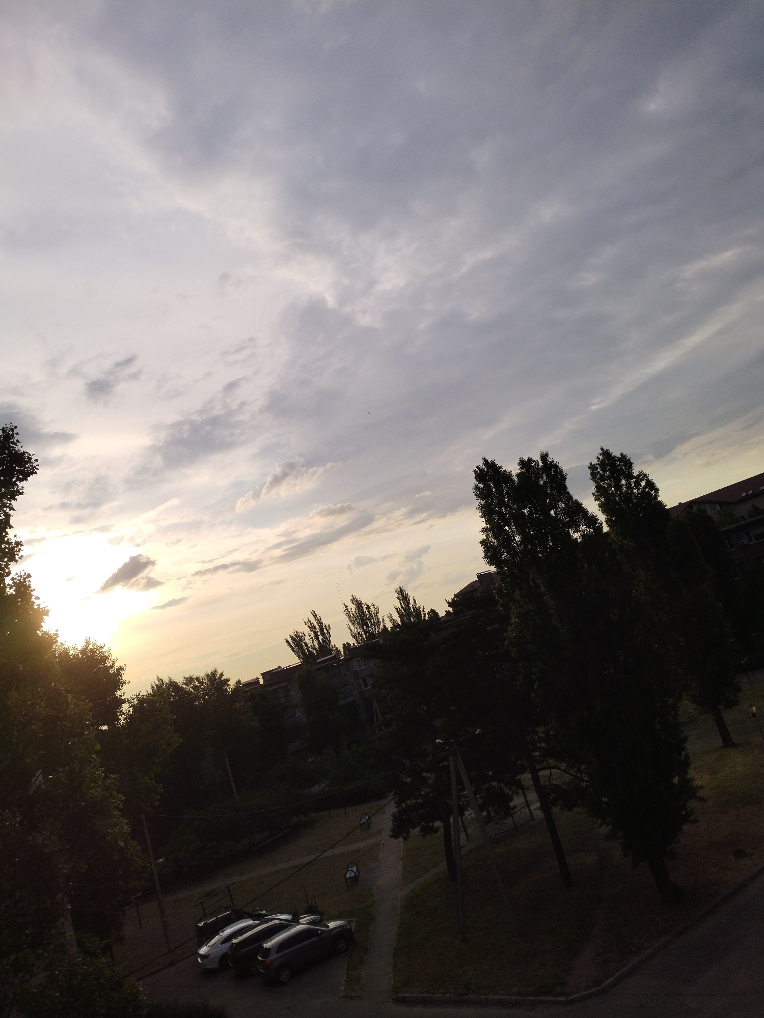 The image depicts an urban residential or park area in Horishni Plavni, Ukraine, during late afternoon or early evening. The sun is low on the horizon, partially obscured by dark foliage on the far left, casting a bright lens flare and illuminating scattered clouds. The sky, occupying the upper two-thirds, transitions from bright white and pale yellow near the sun to shades of grey and muted blue further right. A small dark object, possibly a bird, is visible against the sky in the upper right.
The lower third of the image is dominated by numerous trees, primarily forming dark silhouettes against the brighter sky, indicating backlighting. Several multi-story residential buildings, likely apartment blocks, are visible in the mid-ground, their details obscured by distance, shadow, and trees. One building on the far right has a reddish-brown roof.
At ground level, an asphalt surface, possibly a parking lot or access road, is visible in the lower-middle and lower-right. Three vehicles are parked: a white sedan, a dark-colored sedan, and a dark SUV. Green grassy areas are interspersed with paved pathways. A utility pole with visible lines stands near the parked cars on the left. No individuals are discernible within the scene.