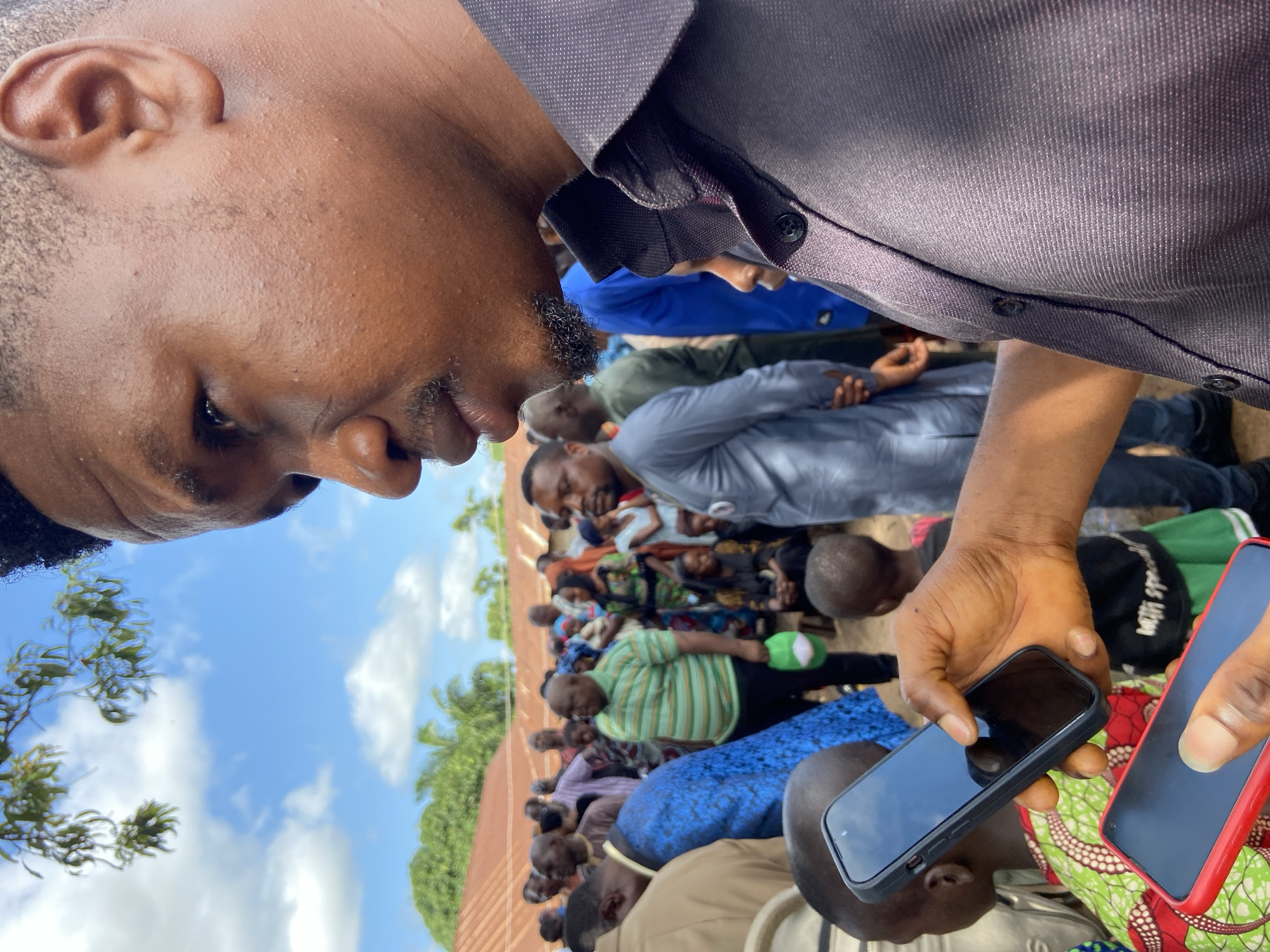 The image captures an outdoor gathering in Mararaba, Nigeria. In the foreground, a man of African descent is seen in a right-profile close-up, positioned on the right side of the frame. He has short, dark, curly hair, a mustache, and a short beard. He is wearing a dark, possibly purplish-grey, long-sleeved collared shirt with visible buttons. His right hand holds a black-cased smartphone vertically with a dark screen, and his left hand, partially visible below, holds a red-cased smartphone horizontally, also with a dark screen. Parts of small children are visible in the immediate foreground, one wearing a green and red patterned garment.
In the midground, a crowd of approximately 20-30 individuals, primarily adults of African descent, is gathered informally. Their attire varies, including a man in a green and white striped polo shirt holding a green cap, a person in a bright blue patterned garment, and another man in a light grey tunic. The individuals are facing various directions within the crowd.
The background features several single-story buildings with reddish-brown corrugated metal roofs. Green trees and dense vegetation are visible around and behind these structures. The sky overhead is bright blue with scattered white cumulus clouds.