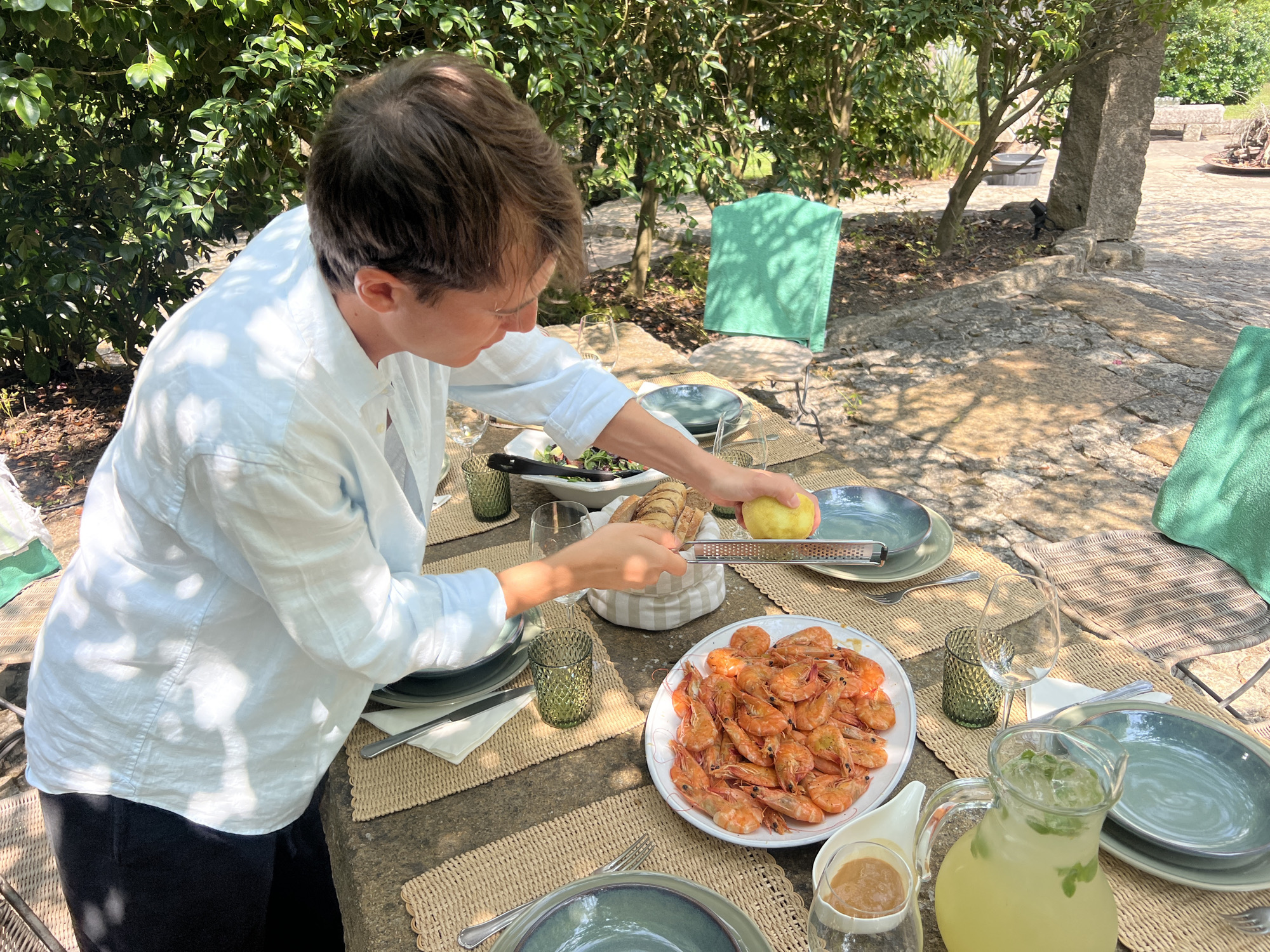 A person is grating a yellow fruit, likely a lemon or apple, into a grater over a table outdoors. The table is set with plates, glasses, cutlery, and a pitcher of lemonade. A large platter of cooked shrimp sits on the table. The background shows lush greenery and stone structures, suggesting a garden setting in Vila Nova de Cerveira, Portugal. Two green towels are draped over chairs.