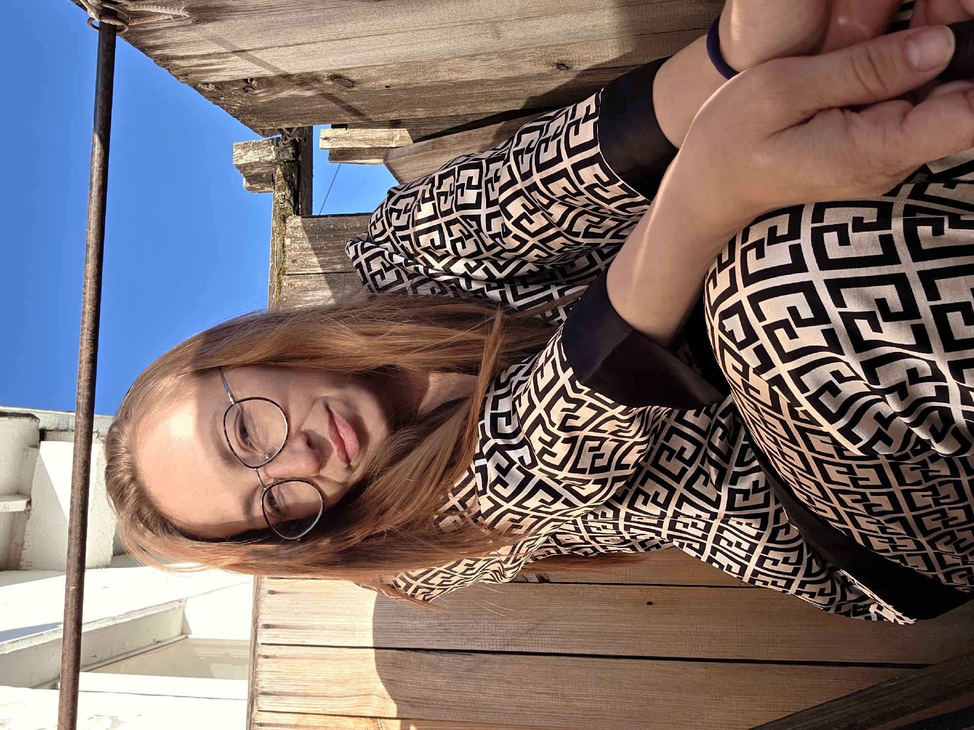 A woman with long auburn hair and round-framed glasses is photographed outdoors, sitting on a wooden platform. She is wearing a black and white patterned top with black accents and has her hands clasped. The background includes a bright blue sky and parts of the wooden structure she is sitting on. The location is potentially the city of Chaikovsky, Russia.