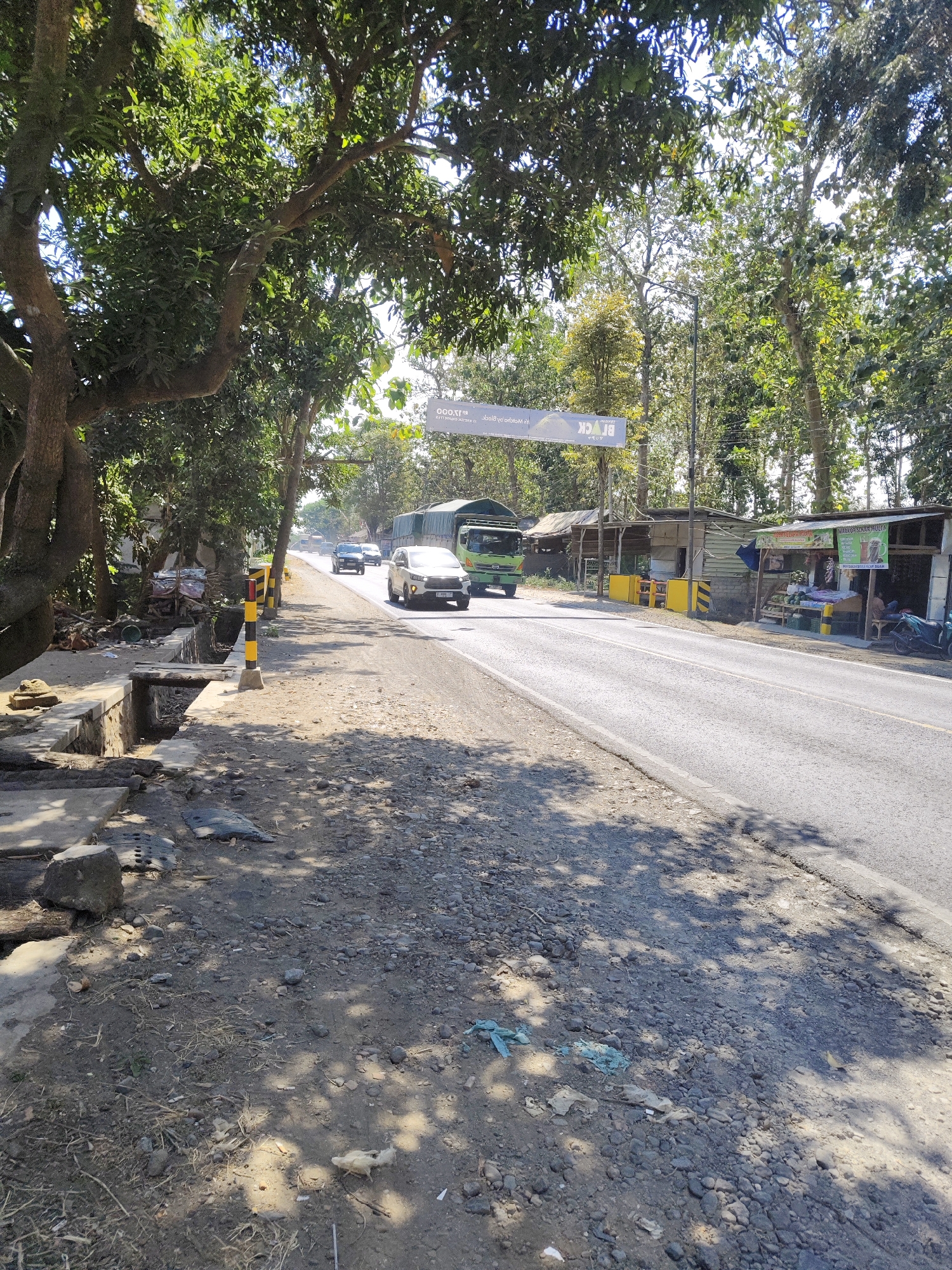 The image shows a road scene in Gambirono, Indonesia. The road is asphalt and has two lanes. On the left side of the road, there is a ditch, some debris, and a yellow and black striped pole. There are also trees with green foliage on the left side, casting shadows on the road. On the right side of the road, there are several small shops with roofs and open fronts, selling various items, including fruits and beverages. A motorcycle is parked in front of one of the shops. A white car and a green truck are driving on the road, along with other vehicles in the distance. A banner is suspended above the road in the distance. The sky is clear and blue.