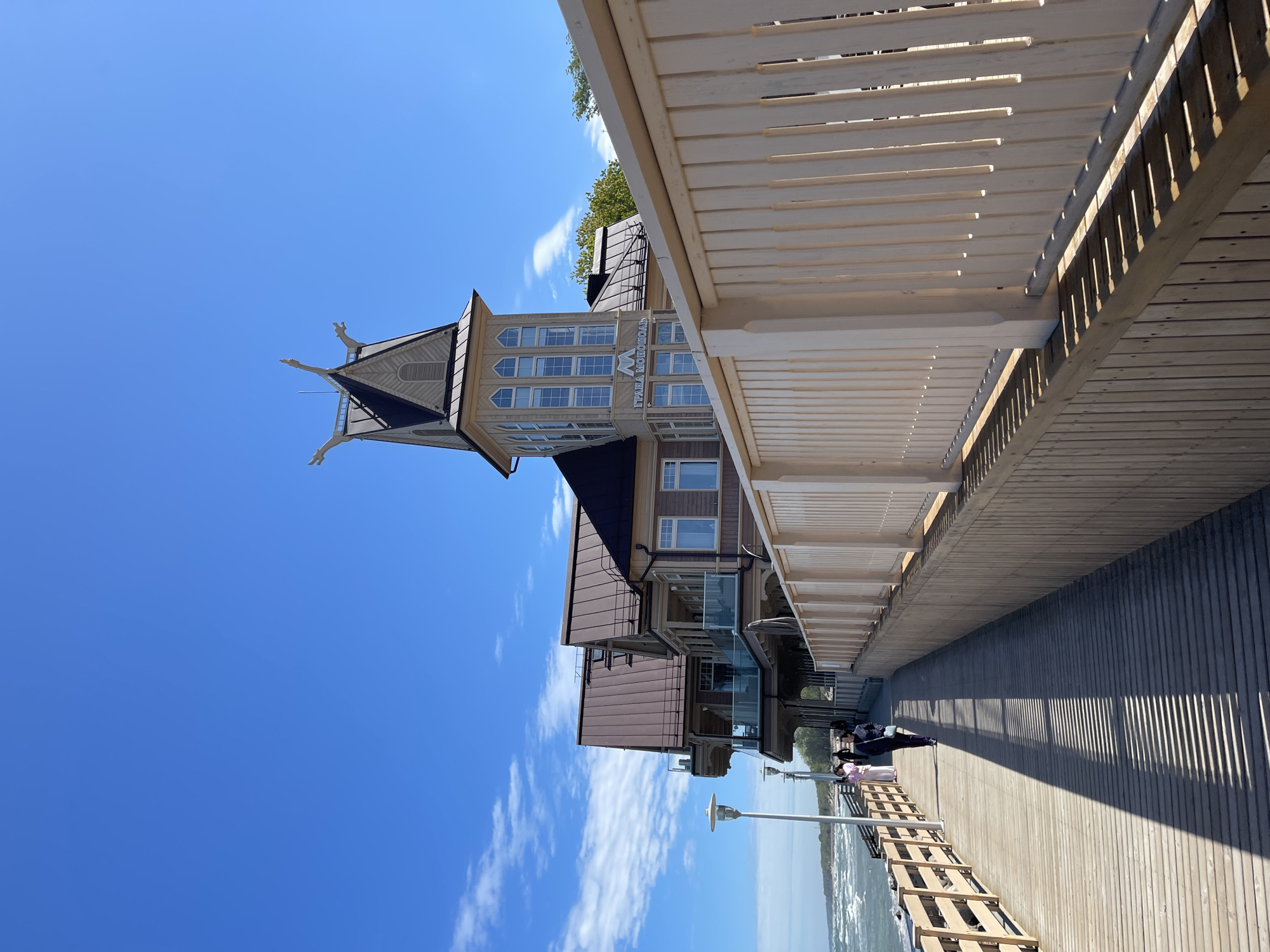The image captures a sunny day in Zelenogradsk, Russia, featuring the "GRAFAS" Hotel, which is a distinctive building with a multi-storied tower. The hotel's architectural details, including decorative elements on its roof, are visible against a bright blue sky with scattered clouds. The photograph is taken from the pier, showing a wooden boardwalk with railings and shadows cast by an overhead structure. People are sitting or walking along the pier, enjoying the view of the water and the cityscape. A row of street lamps stands along the edge of the pier.