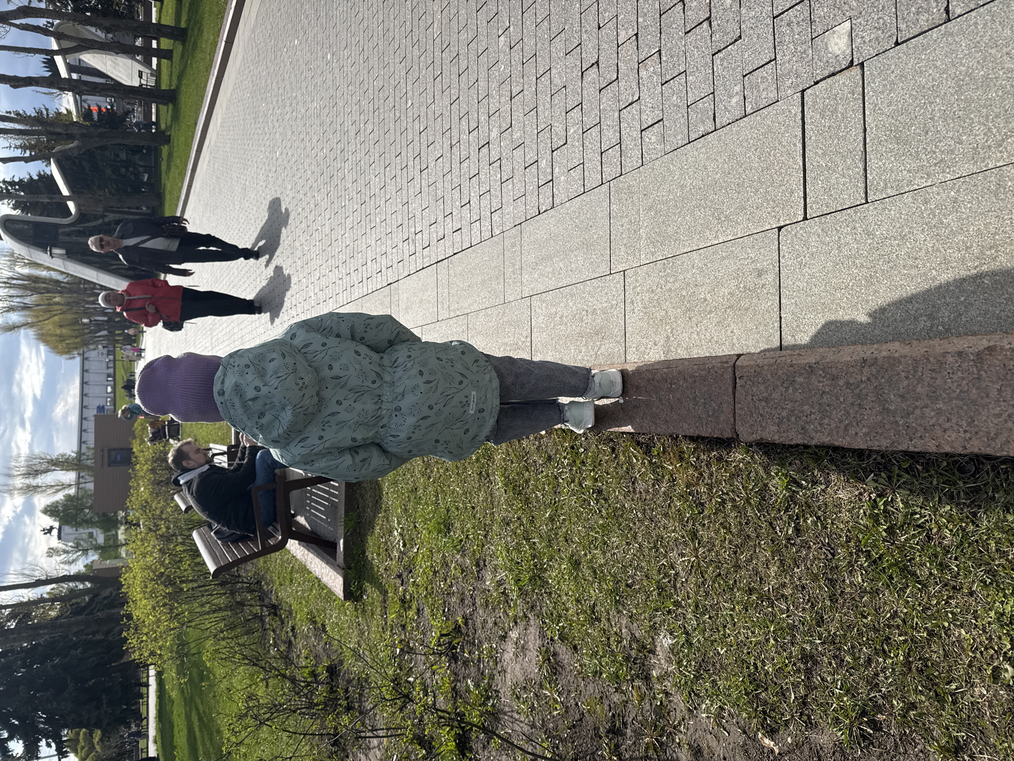 A sideways outdoor image shows a park in what appears to be Moscow, Russia. In the foreground, a child wearing a lilac-colored hat, a light green jacket, gray pants, and white sneakers stands with their back to the viewer. They are standing near the border of a grassy area and a paved walkway. To the child's left, a bench is occupied by a man wearing a dark jacket and jeans. In the background, two adults are walking on the paved walkway, and beyond them, what appears to be a city landscape is visible. The sky is partly cloudy.