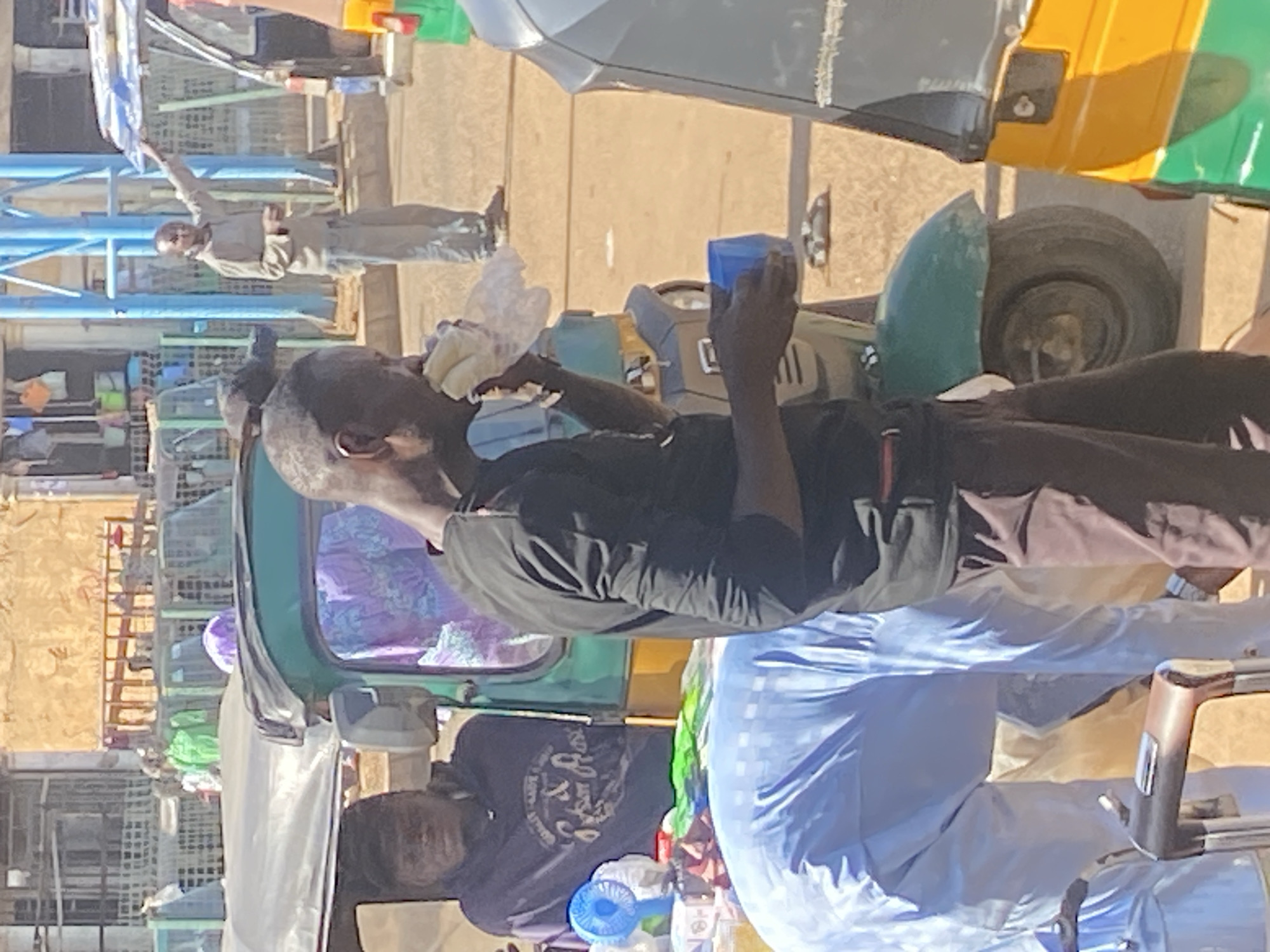 A man is eating in front of a motorized tricycle in a street in Bukuru, Nigeria. The man is wearing a black long-sleeved shirt and brown pants. He is holding food in his right hand and a blue cup in his left hand. The motorized tricycle is green and yellow. In the background, there are other vehicles and people. The street is paved with asphalt. The scene appears to be in a market or a busy street in a city.