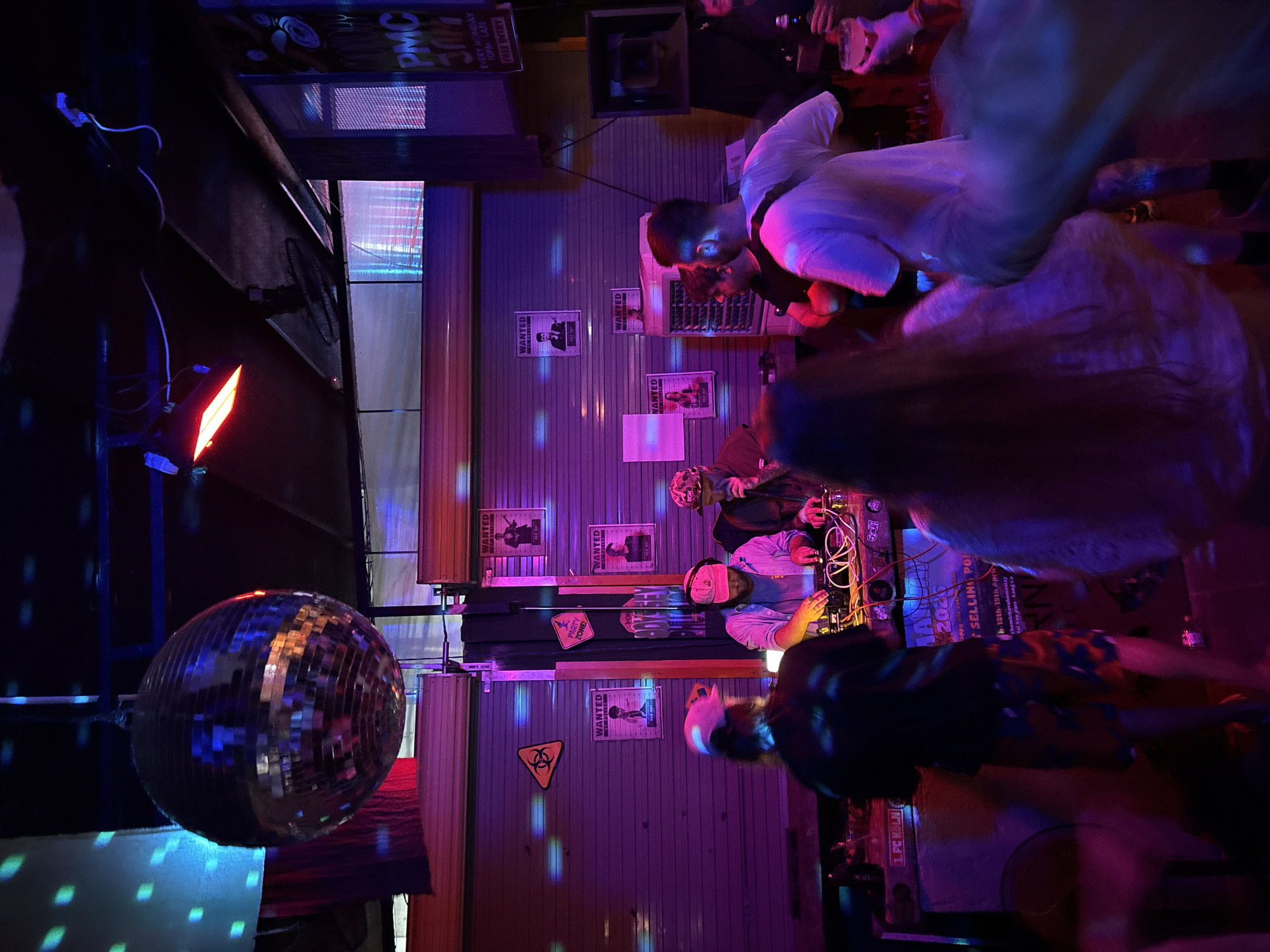 The image captures an energetic indoor scene at what appears to be a bar or club in Đà Nẵng, Vietnam, at night. The setting is dimly lit, dominated by vibrant purple and red artificial lighting that reflects off surfaces, creating a dynamic atmosphere. A large, prominent disco ball hangs in the left foreground, casting scattered light patterns across the room.
Several people are present, indicating a social event or party. In the midground, individuals are gathered around a DJ setup featuring mixing equipment and cables, suggesting a DJ is performing. One person in a white shirt is actively engaged with the console, while others stand nearby or are visible as blurred figures, implying movement and a lively crowd.
The walls, possibly corrugated metal, are adorned with multiple "WANTED" posters featuring stylized caricatures. Other notable signage includes a yellow "PARTY ZONE!" triangle with a biohazard symbol, and a banner promoting an event for "18th-19th April" that mentions "homichavahomestay" and "Tai Van pass-Lang Co." Additional text on signs reads "PMC," "EVERY MONDAY," "10PM LATE," and "FREE ENTRY," suggesting regular events at the venue. A "1. FC KÖLN" sticker is also visible. The combination of lighting, music equipment, and animated people depicts a vibrant and casual nightlife experience.