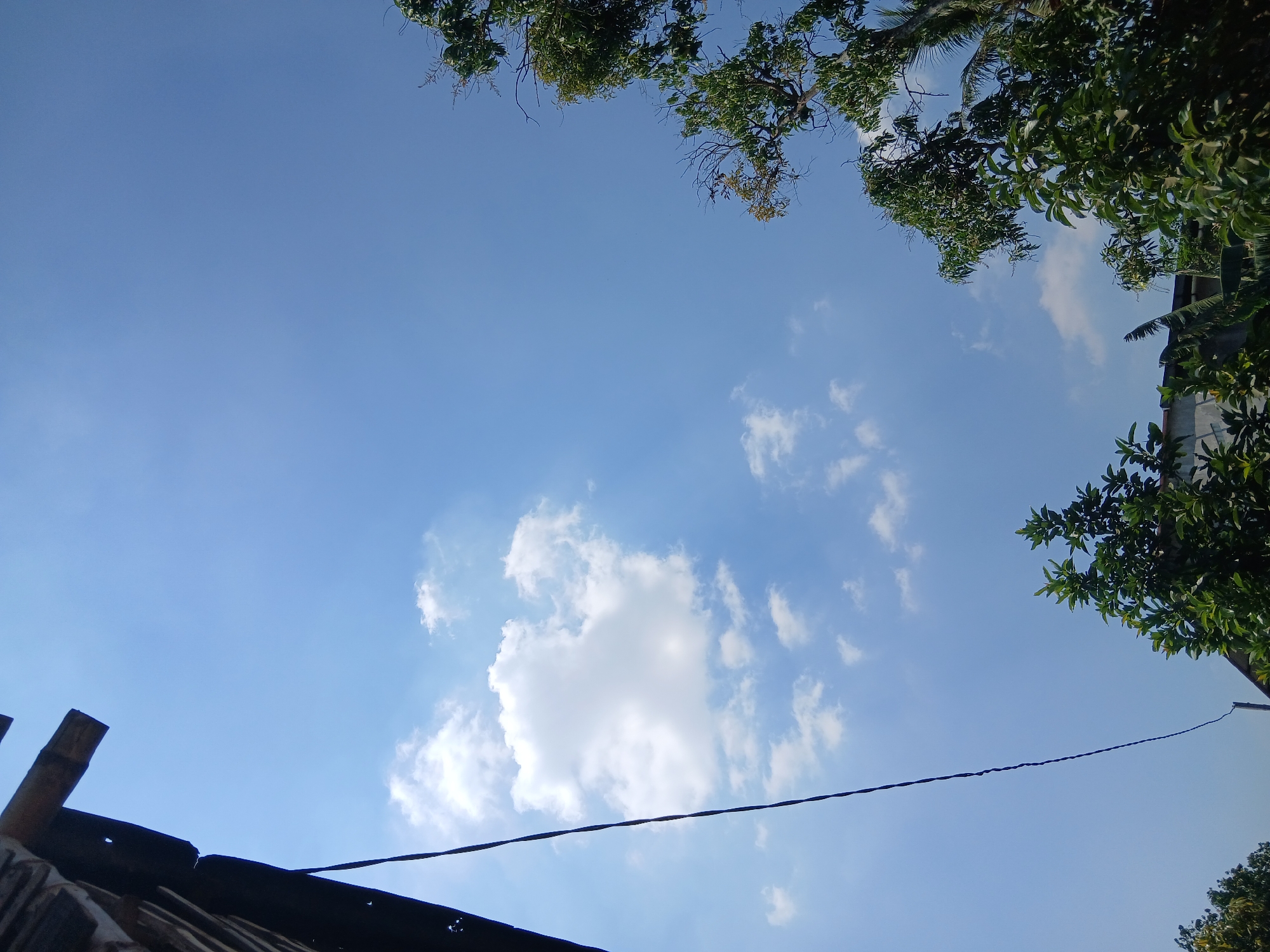 The image depicts an outdoor scene with a bright blue sky. In the upper left corner, a dark, weathered structure, possibly a roof edge with visible wood and corrugated material, juts into the frame. A dark wire hangs diagonally from the top left towards the center of the image. Scattered white clouds dot the sky, with a larger, more prominent cloud in the upper left quadrant. The lower portion of the image is dominated by the lush green foliage of trees and some roofing structure, suggesting a residential or semi-rural environment. The overall impression is of a clear day in Kademangan Kulon, Indonesia, with elements of the built environment intertwined with nature. There are no people or animals visible in the scene.