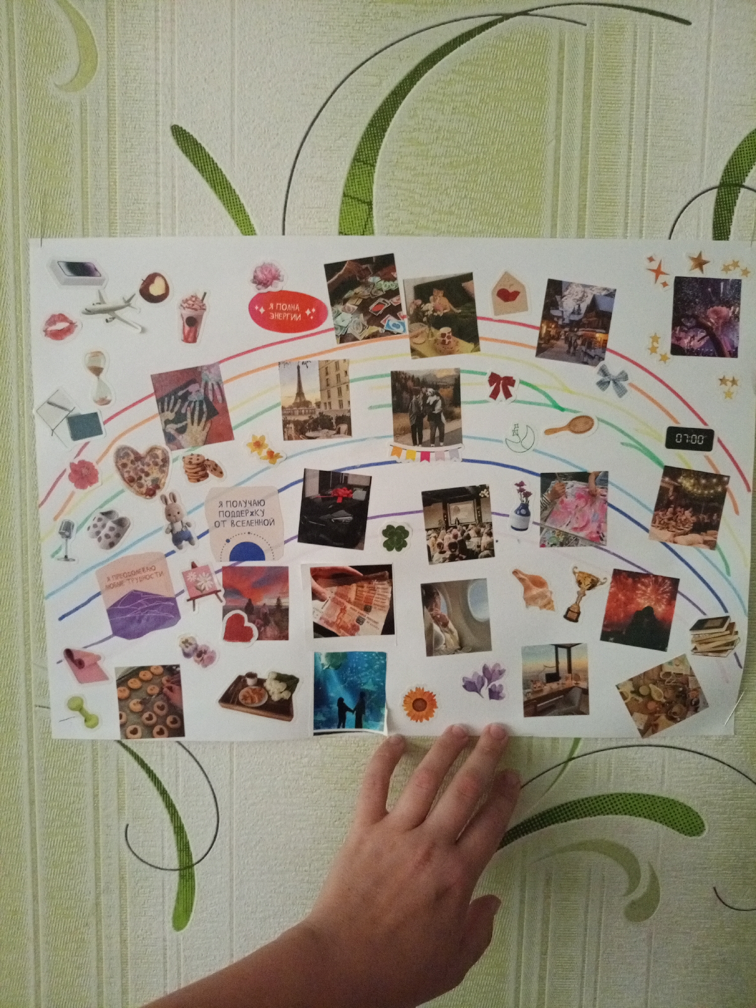 The image shows a collage of photographs and stickers adhered to a wall with light green striped wallpaper. The collage is arranged in a rainbow-like arc across a white sheet of paper. Various small photographs depict scenes such as a cityscape with the Eiffel Tower, people gathered at an event, a trophy, a plane interior, and food. There are also numerous stickers, including lips, a coffee cup, a heart, a bunny, flowers, a microphone, and text in Russian. A hand is visible in the lower right corner, reaching towards the collage. The environment appears to be indoors, likely a room in a residential setting, given the wallpaper and personal nature of the collage. The lighting is diffuse, suggesting ambient light from an unseen source.