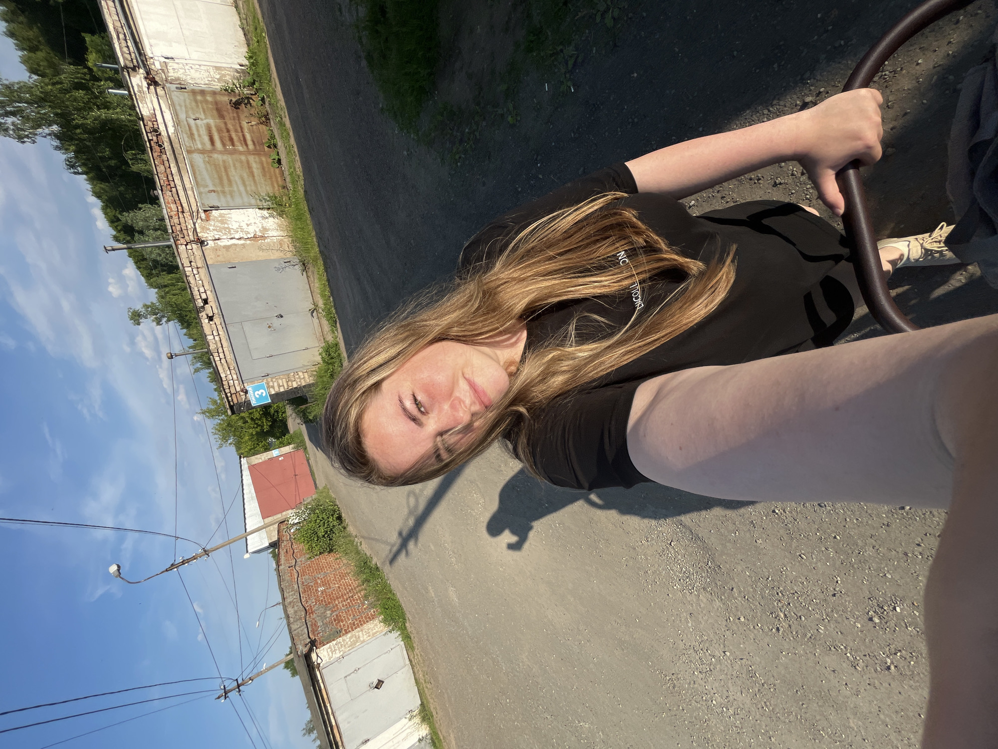 A woman is taking a selfie outdoors in Chaikovsky, Russia, on a sunny day. She is standing on a dirt road with garages lining both sides. She is wearing a black t-shirt and sneakers. In the foreground, her arm is extended holding a stroller. The garages are made of brick and metal, some with red doors. There are power lines and streetlights visible in the background. The sky is blue with some clouds.