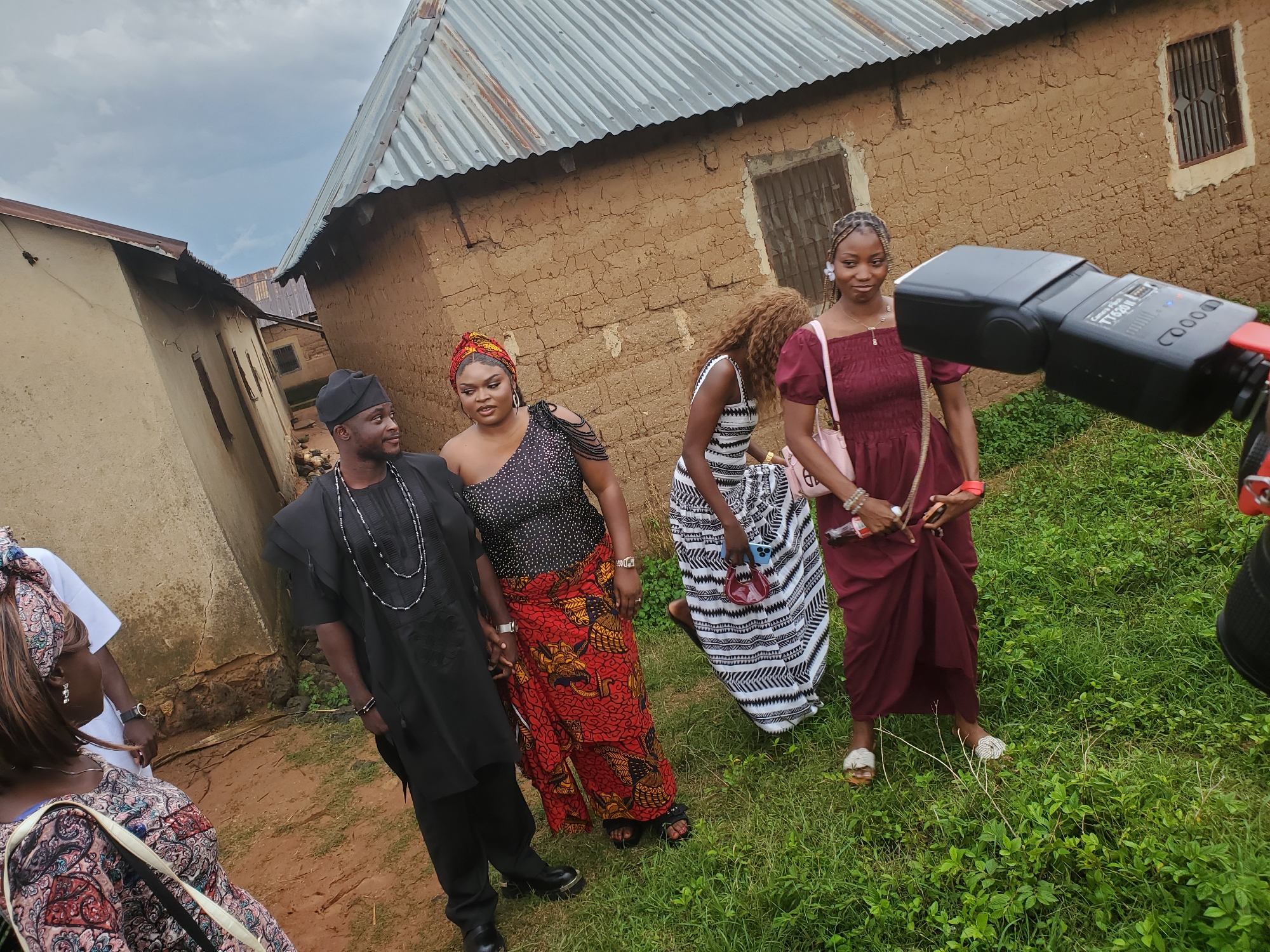 A medium shot captures a group of people in an outdoor setting, likely within the city of Jos, Nigeria, characterized by the presence of traditional mud-brick buildings with corrugated iron roofs. The foreground is dominated by lush green grass.
In the center of the frame, a man dressed in a black traditional outfit and a black cap stands facing left, with a beaded necklace around his neck. To his right, a woman in a black top adorned with rhinestones and a vibrant red and yellow patterned skirt, also wearing a matching headscarf, turns her head to look towards the man. Behind them, another woman in a white and black striped dress is visible, holding a small red purse and a phone. Further to the right, a woman in a maroon dress with puffed sleeves and sandals stands, her body turned towards the viewer.
In the extreme right foreground, a black camera flash unit is prominently displayed, indicating a potential photography or videography event. The background shows the rough texture of mud-brick walls and the aged appearance of the corrugated metal roofing, suggesting a rural or older residential area within Jos. The sky is overcast.