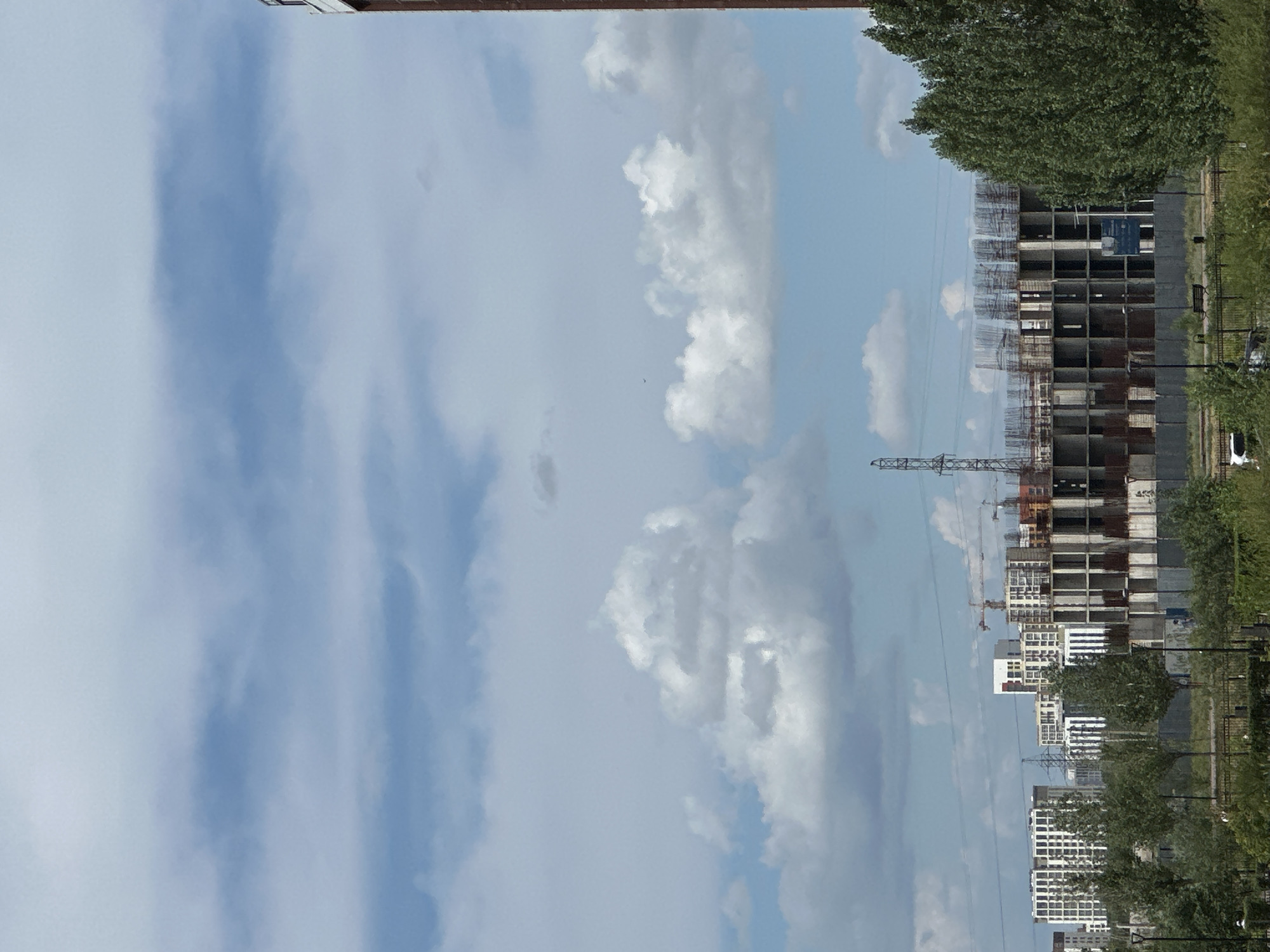 The image shows a cityscape with several construction sites. In the foreground, there are trees and a fence. Beyond that, there are various buildings under construction, some with exposed rebar. In the background, more completed buildings are visible, along with power lines and a transmission tower. The sky is partly cloudy, with a mix of blue sky and white clouds. There are no people clearly visible in the image. The scene appears to be an urban or suburban area undergoing development.