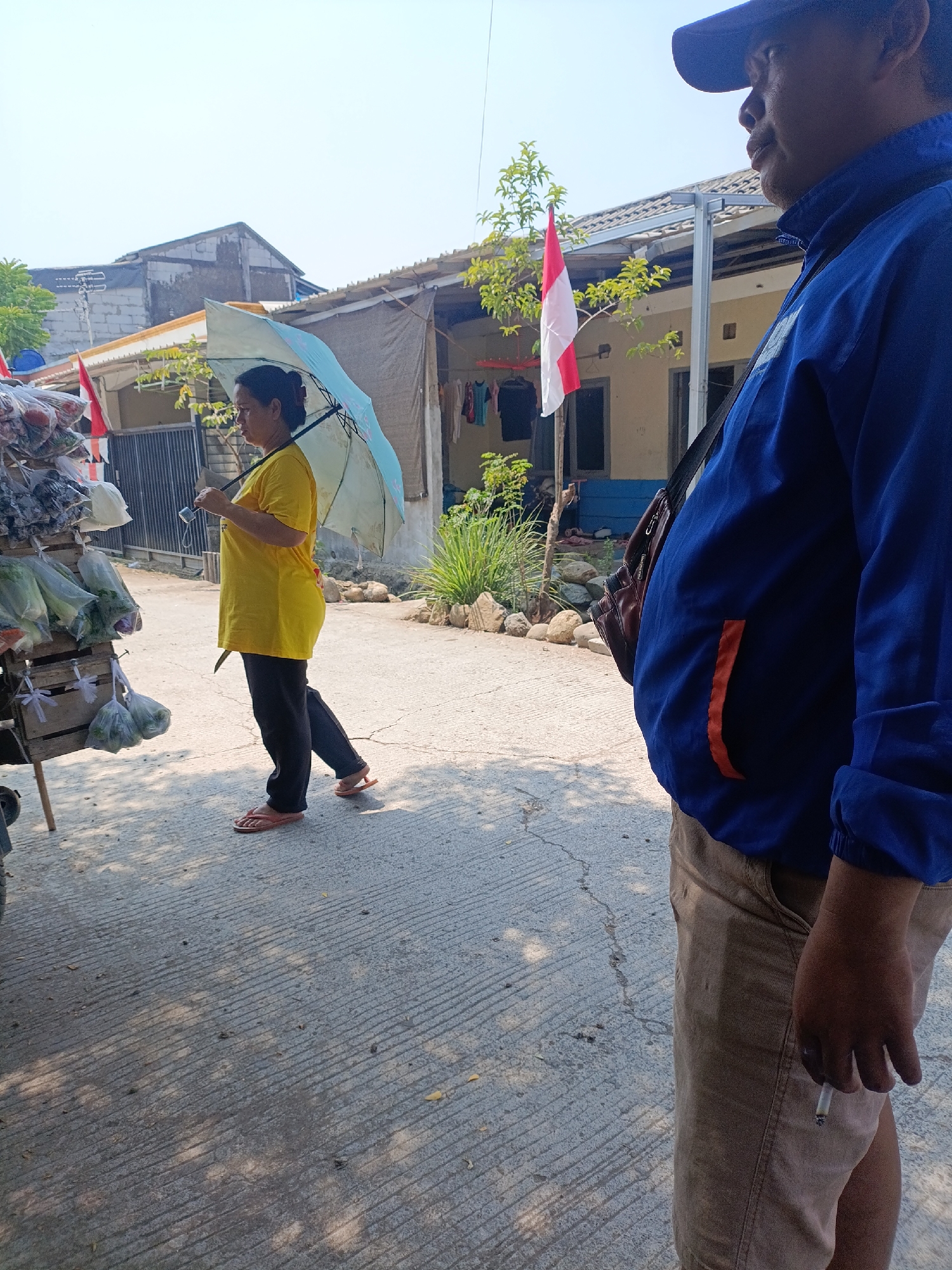 The image depicts an outdoor scene in Cariu, Indonesia, likely on a street or market area. In the foreground, a person wearing a blue jacket, a blue cap, and tan pants is partially visible on the right side, holding a lit cigarette. A brown shoulder bag is slung across their chest. To the left of the frame, a woman wearing a yellow t-shirt and black pants, with orange flip-flops, stands holding a light blue umbrella. She appears to be near a stall displaying various items wrapped in plastic bags, possibly produce. In the background, a red and white Indonesian flag is visible on a pole, attached to a building. The ground is paved with a textured surface, and shadows from trees are cast across it. Buildings and vegetation are visible behind the stall and the flag. The sky is clear and bright, suggesting a sunny day.