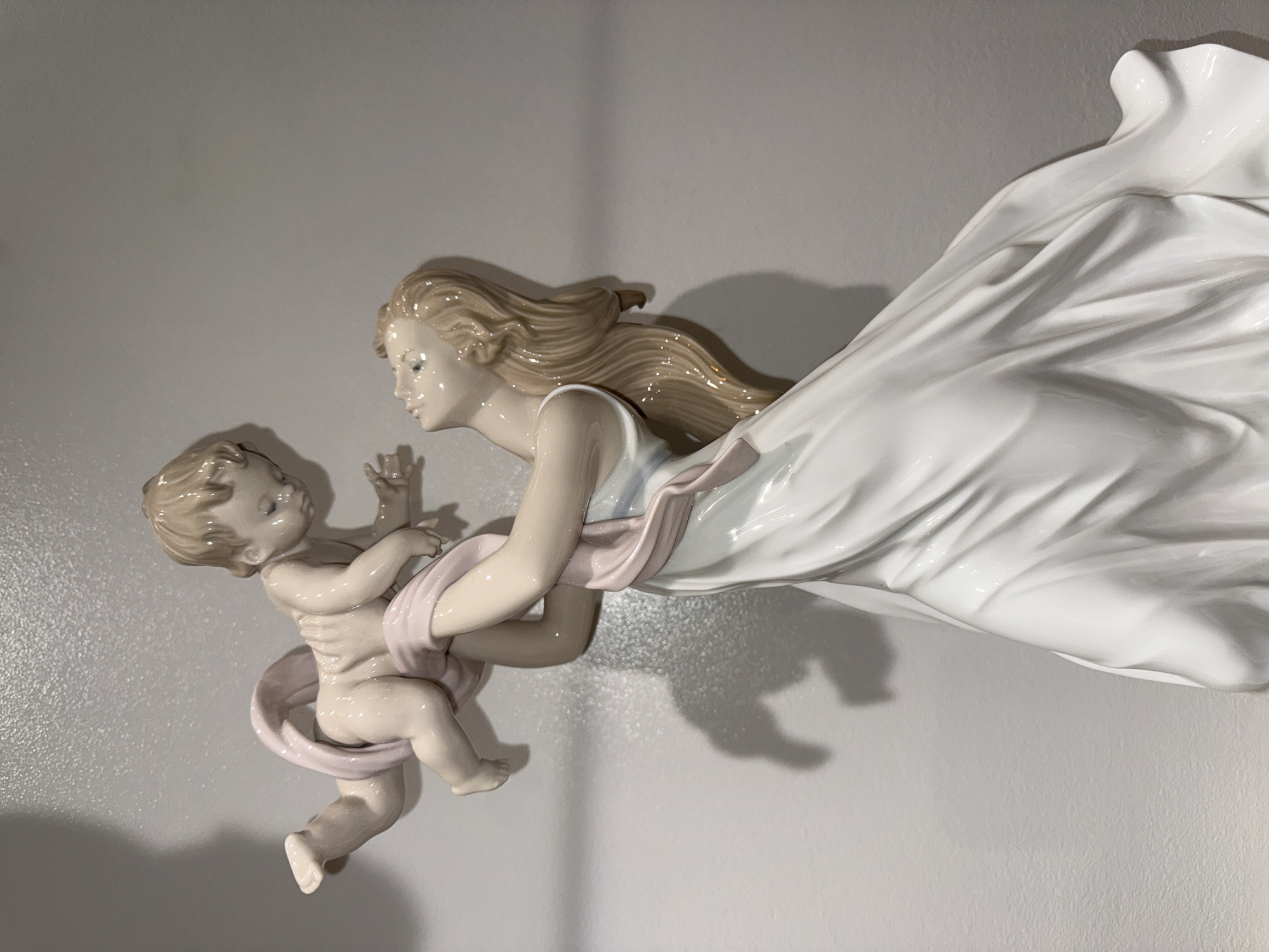 A glossy porcelain figurine group is centrally depicted against a textured, light grey wall in Barcelona, Spain. The sculpture portrays an adult female figure in a long, flowing white gown with a pink sash draped across her body and the infant she is holding. Her head is tilted sharply upward, gazing at the baby, with long, light brown hair styled to flow down her back. The nude infant, partially enveloped by the pink sash, is held aloft, with its arms reaching towards the woman's face and its legs bent. Both figures possess light beige skin tones and are highly reflective due to the porcelain finish. A distinct light source from the upper left creates strong shadows of the figures on the background wall.