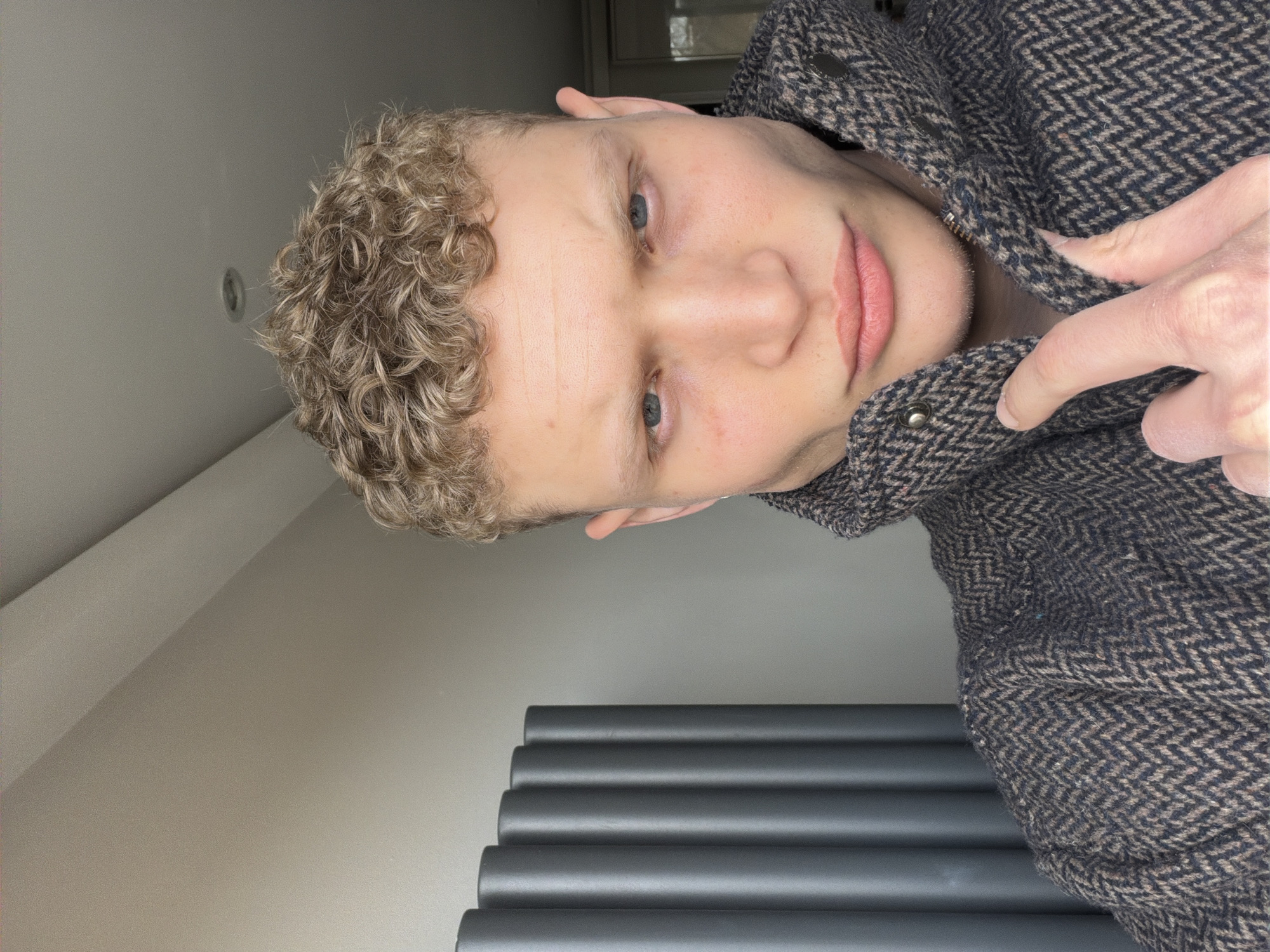 A young person with light, curly blonde or golden-brown hair and striking blue eyes is depicted, looking directly at the camera with a subtle, calm expression and slightly parted lips. They appear to have fair skin.
The individual is wearing a dark, textured jacket with a distinct herringbone pattern in shades of brown and beige/cream. Their right hand is visible, gently holding and adjusting the collar or lapel of the jacket, near a metallic button snap.
The setting is indoors, with plain, light-colored walls and ceiling serving as the background. To the lower right of the frame, a modern dark grey radiator with prominent horizontal, cylindrical heating elements is visible. A small, circular recessed light fixture can be seen on the ceiling above the person's head, towards the left.
The photograph is taken at a significant tilt, giving the impression that the subject is lying down or the camera was held at an angle. The lighting is soft and even, illuminating the subject's face without harsh shadows. There is no visible text in the image.