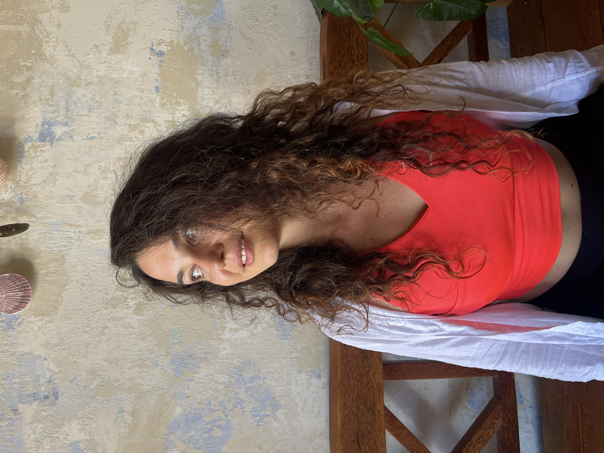 In Phu Thuy Ward, Vietnam, a woman with long, curly, dark brown hair is pictured sitting on a wooden bench. She is wearing a bright orange tank top and an unbuttoned white shirt over it. The background features a textured wall with patches of blue and cream paint, and two decorative seashells are affixed to the upper portion of the wall. To the right of the woman, a lush green plant is visible, partially obscuring a wooden support structure. The woman is looking to her left, with a subtle smile on her face. The lighting suggests an indoor setting.
