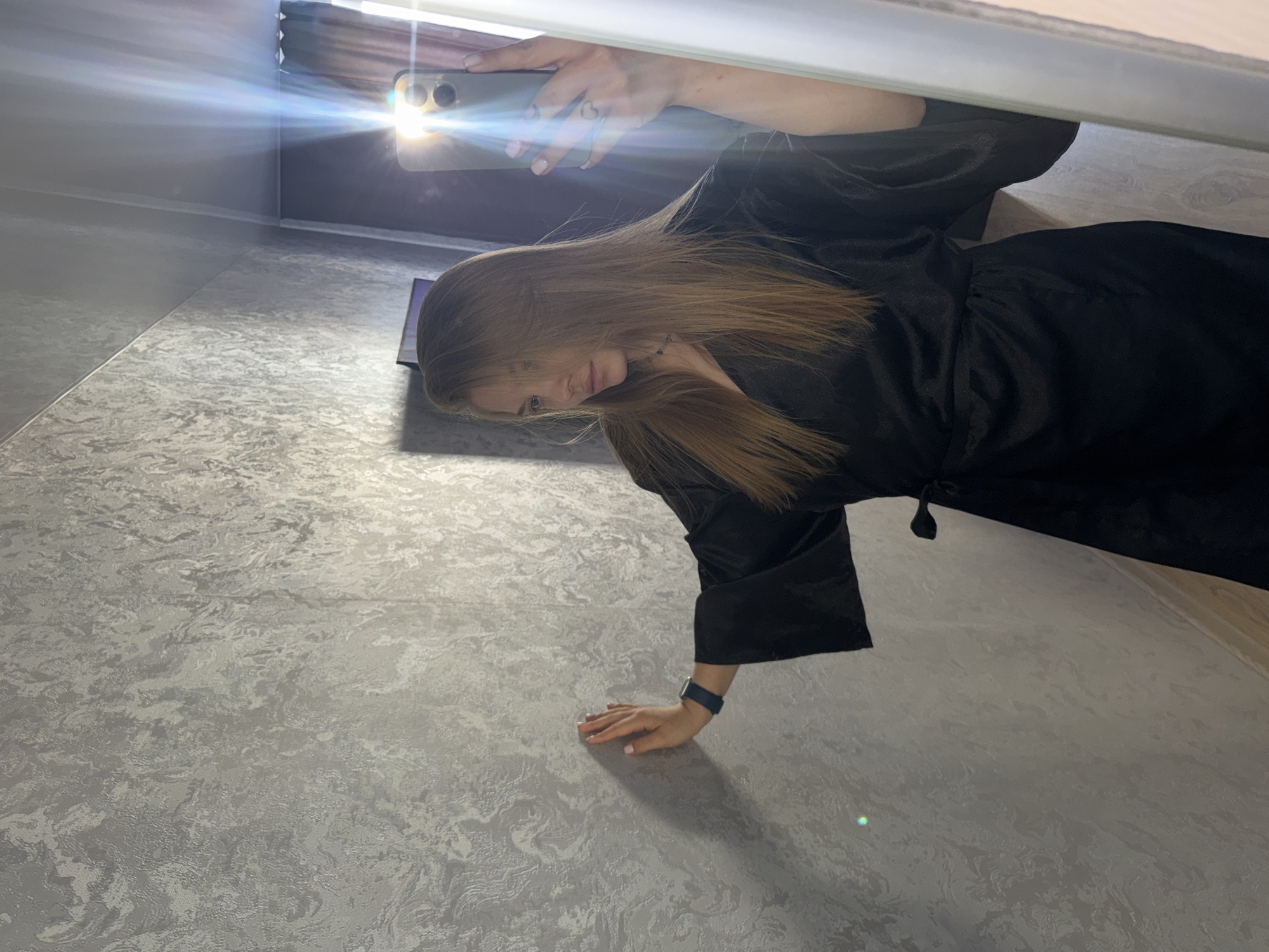 A person is seen taking a selfie in what appears to be an indoor setting. The person has long, light brown hair and is wearing a black satin robe. They are holding a smartphone with the camera facing them, and the flash is on. Part of a window with blinds is visible in the upper background. The floor has a light grey patterned surface. The location is Chaikovsky, Russia.
