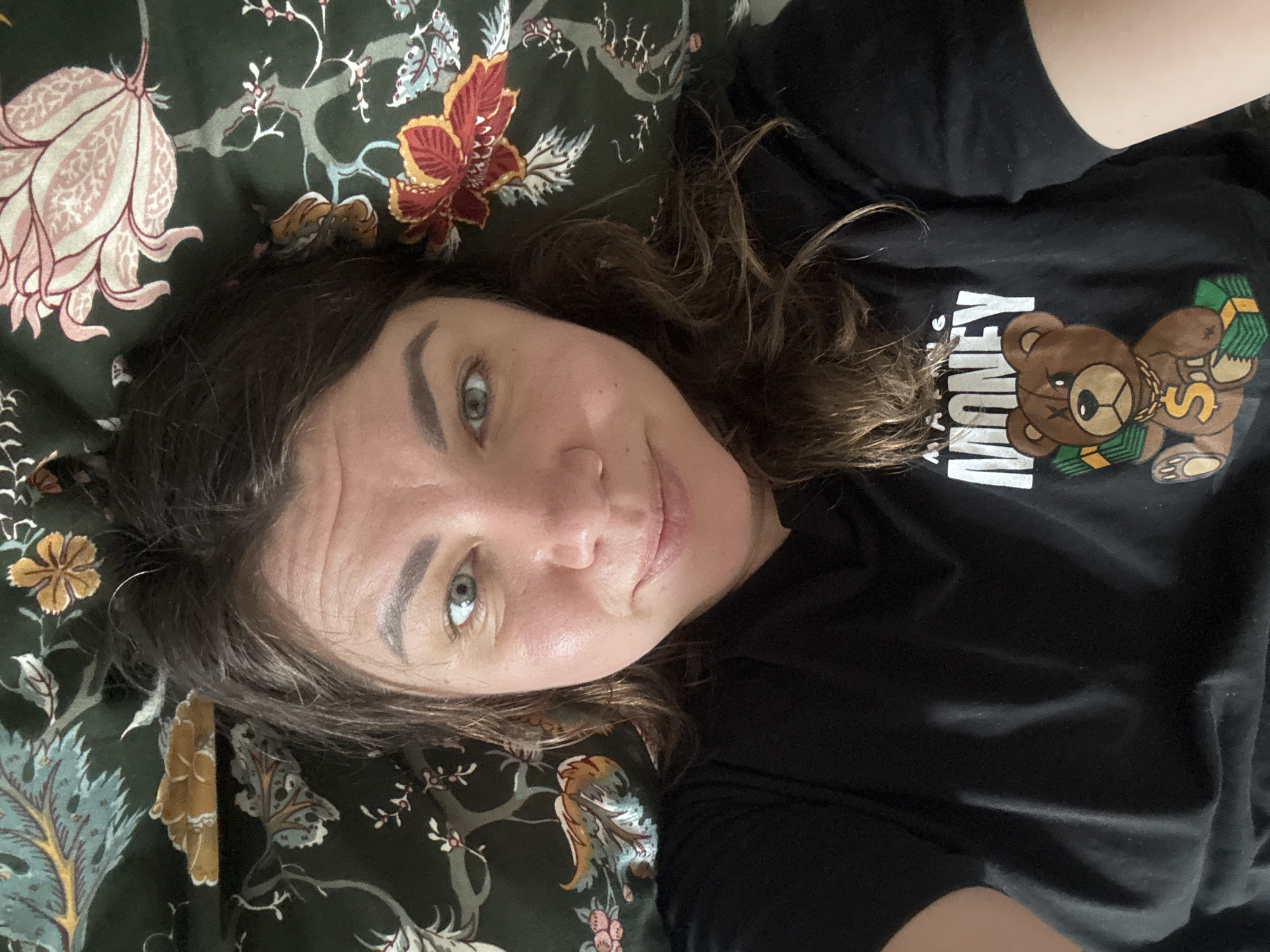 A woman with brown hair and green eyes is lying on a dark green floral-patterned surface, possibly a bed or sofa. She is wearing a black t-shirt with a graphic print of a teddy bear holding money, with the word "MONEY" written in large white letters below it. The teddy bear is brown with a gold chain and a dollar sign on its chest, and it is surrounded by stacks of money. The floral pattern on the surface features large pink and red flowers with some lighter colored leaves and branches. The lighting suggests it could be daytime, possibly indoors, as there are no obvious external light sources or weather cues. The woman's expression is relaxed, with a slight smile. The image appears to be a selfie, taken from a high angle looking down at the woman. The location context provided is Höhenkirchen-Siegertsbrunn, Germany, but there are no specific visual cues in the image to confirm this.