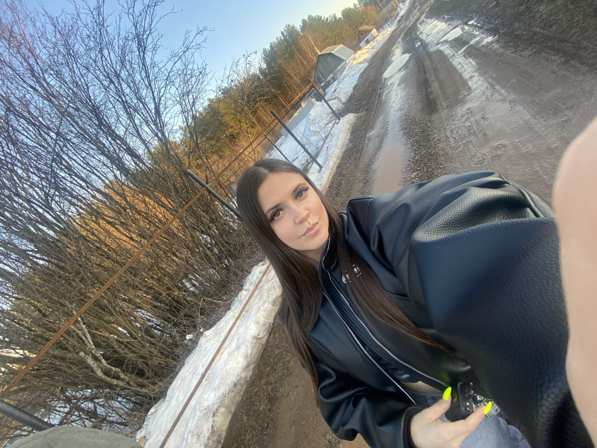 A selfie-style photograph captures a young woman outdoors in what appears to be a rural or semi-rural setting during late winter or early spring. The woman, positioned in the foreground and slightly to the right, has long, straight dark hair and is wearing a black faux leather jacket with visible zippers. She is looking directly at the camera with a neutral to slightly serene expression, her makeup including defined eyebrows, eyeliner, and subtle lip color. Her right hand is visible, holding a small, indistinct object, and her fingernails are painted a bright, neon yellow-green.
The background reveals a landscape indicative of a thaw. To the left, a dense thicket of bare deciduous tree branches fills a large portion of the frame, suggesting trees without leaves. Along the ground, patches of melting snow are visible, bordering a muddy, unpaved road or path that stretches into the distance. This road is marked with tire tracks and numerous puddles reflecting the sky.
Further back, a line of coniferous trees (pines) forms a forest. Beyond the forest, faint structures, possibly old wooden buildings or sheds, can be seen under a clear, pale blue sky. The lighting suggests either early morning or late afternoon, with a low sun casting a gentle, warm light on the distant trees and creating soft shadows. A metal fence or railing is partially visible along the edge of the path near the woman. The overall scene suggests a cold but sunny day, with the remnants of winter still present. No text is visible in the image.