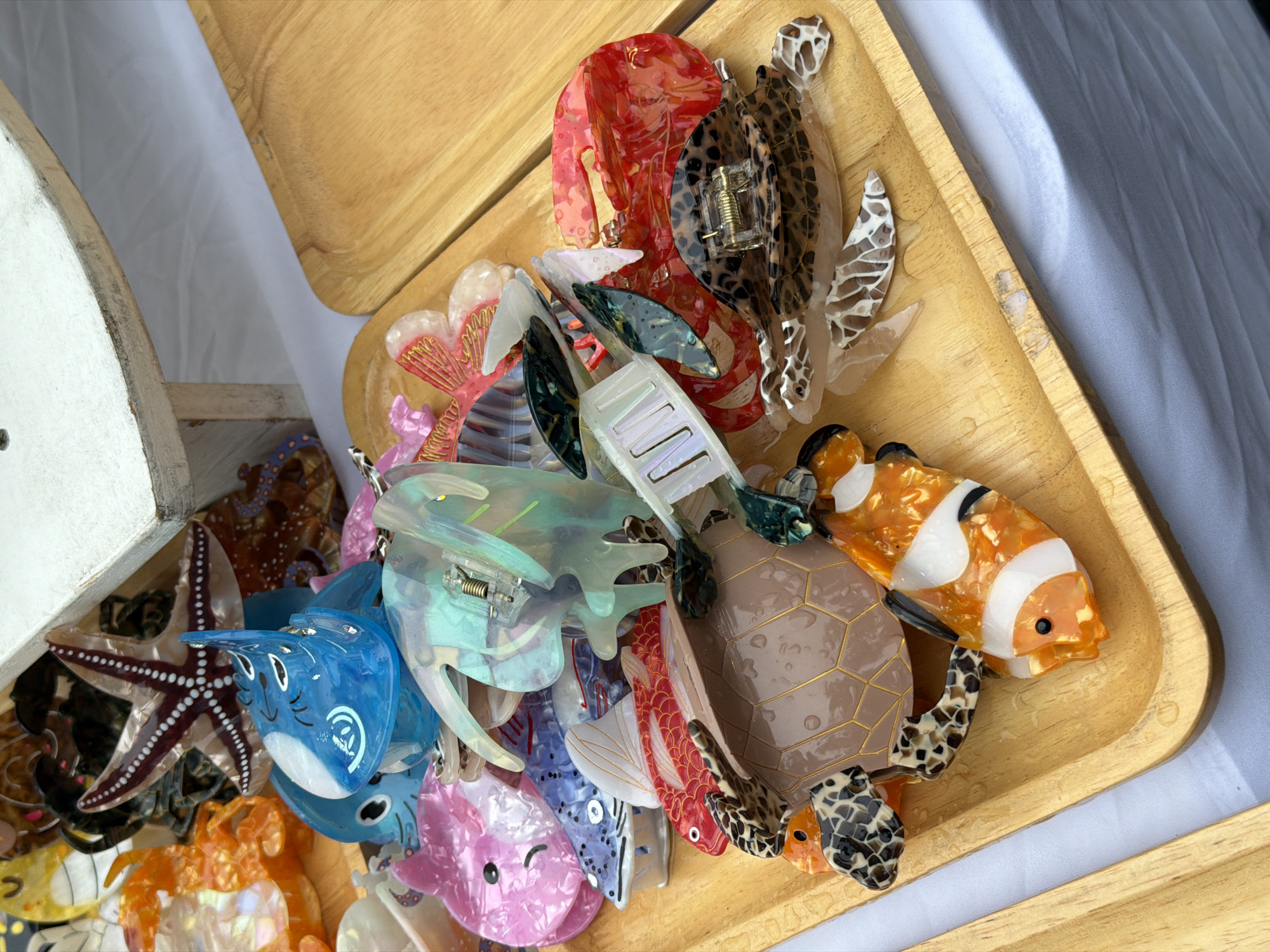 A wooden tray overflowing with colorful acrylic hair claw clips, predominantly shaped like marine life, sits on a white surface. The clips include a blue dolphin, a pink dolphin, a fish with red and white fins, a leopard-print turtle, a clownfish, and various translucent, iridescent clips in shades of green and blue. Some clips are stacked atop a larger, detailed turtle shell clip. In the background, a wooden stand supports an arrangement of faux starfish and other decorative sea creatures, also appearing to be made of acrylic. The overall scene suggests a retail display or a collection of novelty items. The location is identified as being in Choeng Thale, Thailand.