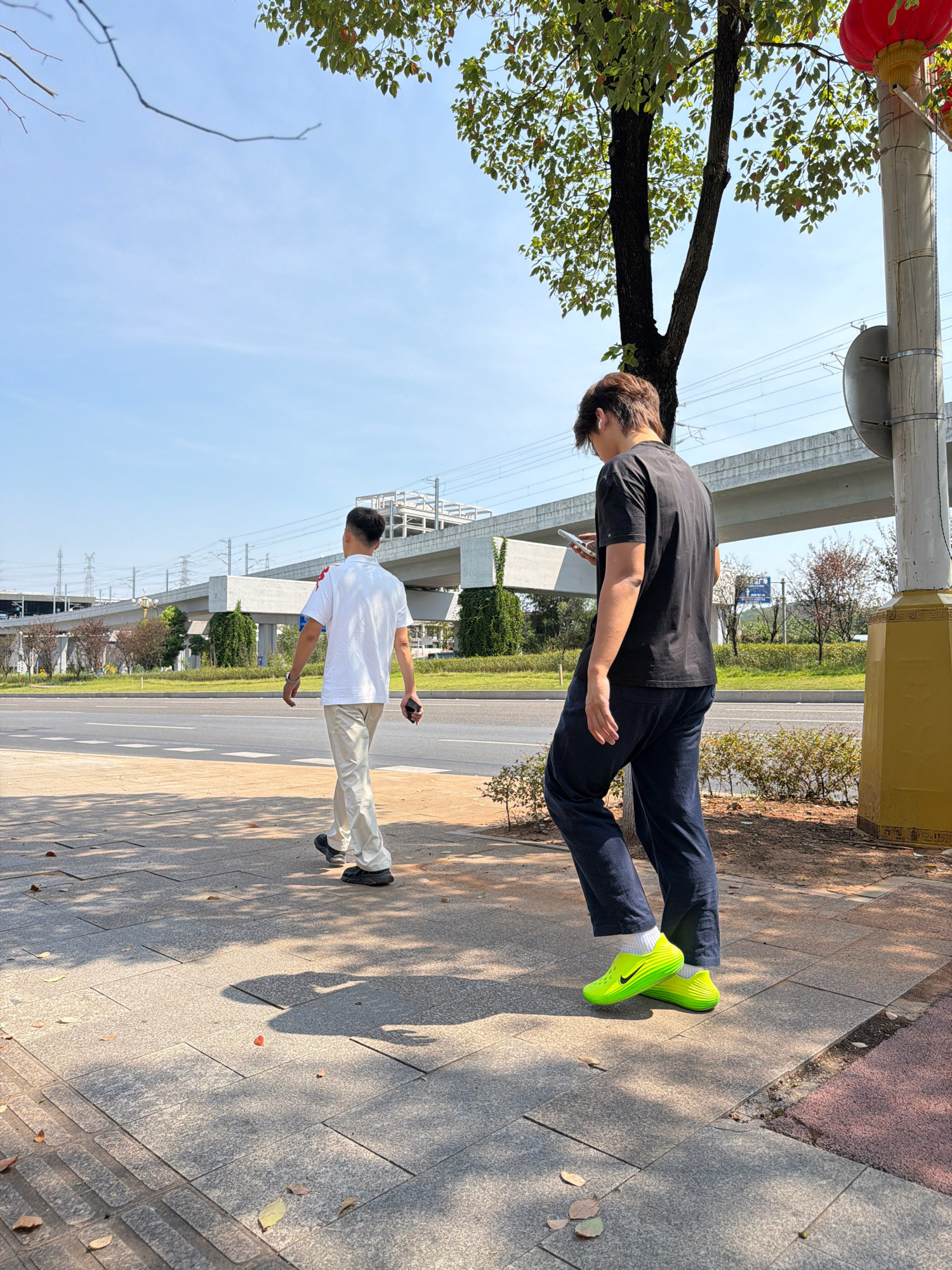 Two young men are walking on a sidewalk in Jiangjin, China. The man on the left is wearing a white polo shirt and beige pants, and he is looking to his left. The man on the right is wearing a black t-shirt and dark blue pants, and he is looking at his phone. Both men are wearing casual shoes. They are walking in the same direction, away from the camera.
The sidewalk is paved with grey tiles and is lined with trees. In the background, there is a road with cars, an elevated train track, and buildings. The sky is blue and clear, with some wispy clouds. The sun is shining, casting shadows on the sidewalk. A red lantern is hanging from a pole on the right side of the image, indicating a festive or traditional element.