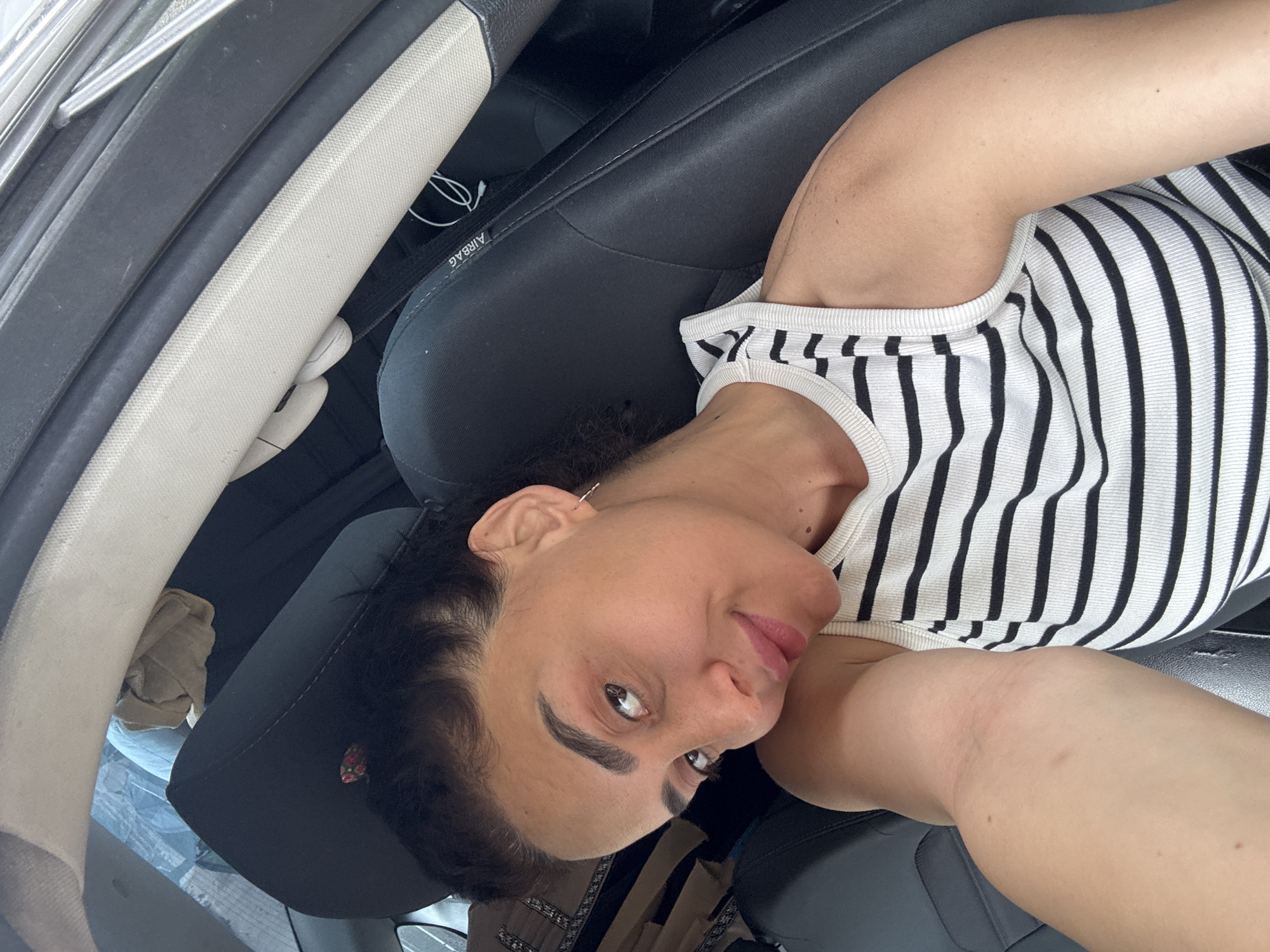 A woman is sitting in the passenger seat of a car, with her head tilted and looking at the camera. She has dark hair pulled back and is wearing a white and black striped tank top. Her left arm is resting on the back of her head, and her right arm is visible from the shoulder down, resting on her lap. The interior of the car, including the black leather seat with an "AIRBAG" label, is visible. The background shows the outside through the car window, suggesting an urban environment, possibly Tbilisi, Georgia, based on the context provided.