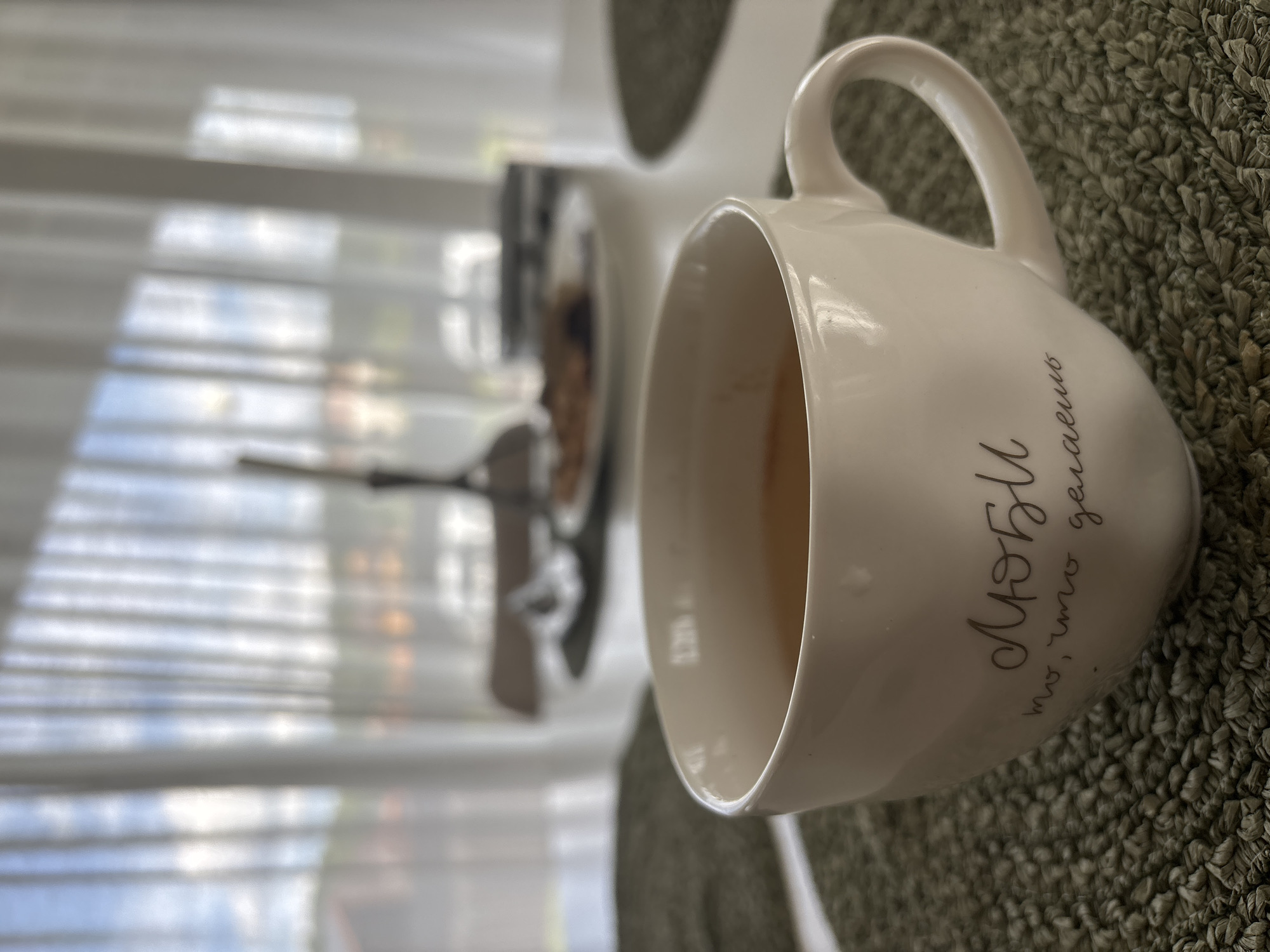 A close-up, low-angle shot captures a white ceramic mug filled with a light brown beverage, likely tea or coffee. The mug rests on a textured green placemat. The mug has cursive writing in a dark gray color that reads "Люби то, что делаешь" which translates to "Love what you do". The background is blurred, showing sheer white curtains filtering natural light, suggesting an indoor setting by a window. Through the curtains, a hint of greenery and buildings is visible, implying a suburban or semi-rural environment. In the far background, a blurred plate with food items and a decorative Eiffel Tower figurine can be discerned. The overall scene evokes a sense of a peaceful morning or afternoon break. The image was taken in Zvenigorod, Russia.