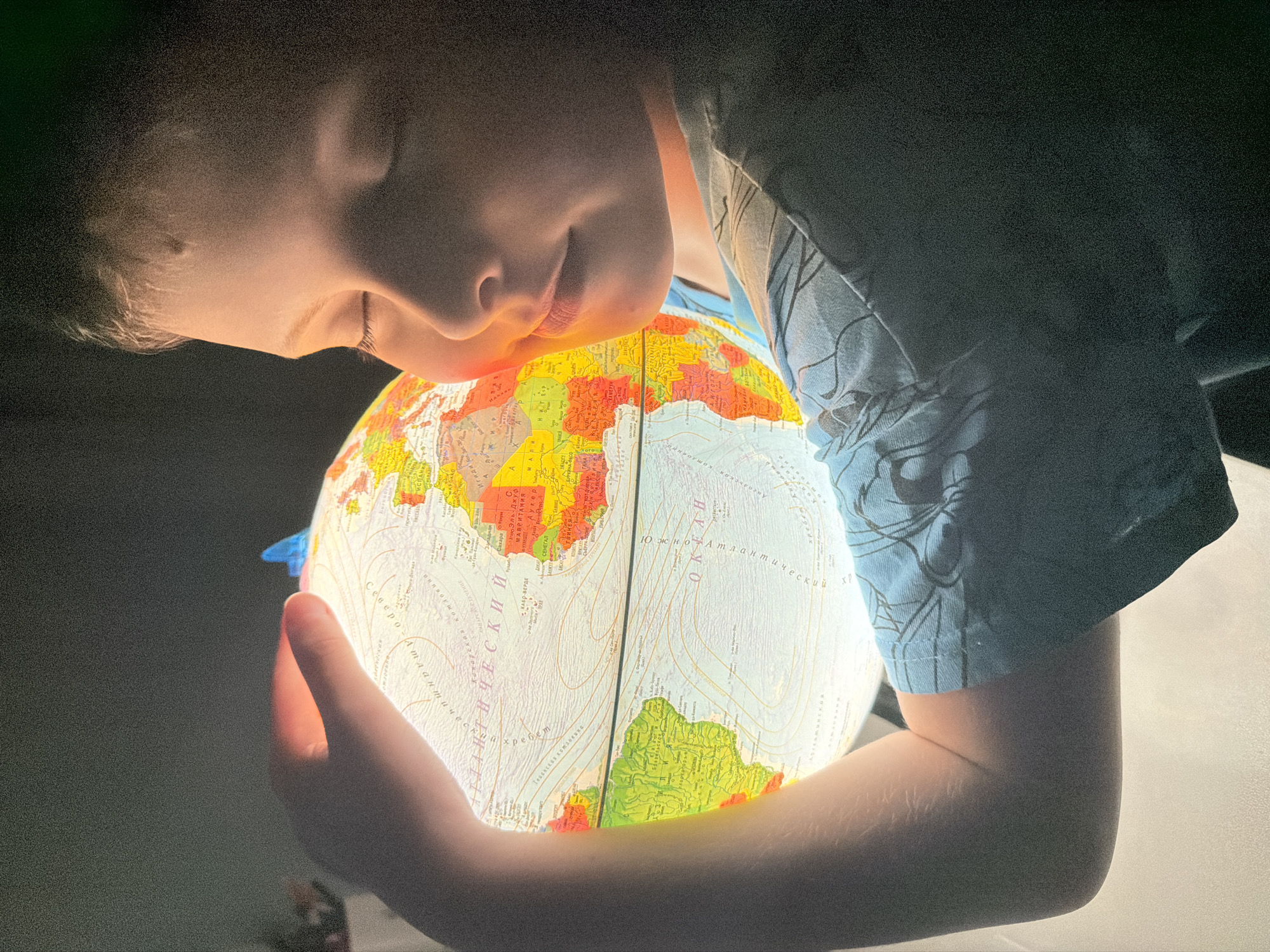 A young person is embracing an illuminated globe with their eyes closed. Their face is close to the globe, which is bright enough to cast light on their features. The globe displays a world map with visible text in Russian, denoting countries and geographical features. The individual is wearing a short-sleeved shirt. The scene is dimly lit, and the background is dark, creating a focused and intimate atmosphere around the subject and the globe. There is no location data available, as the city of Votkinsk, Russia, is not evident in the image.