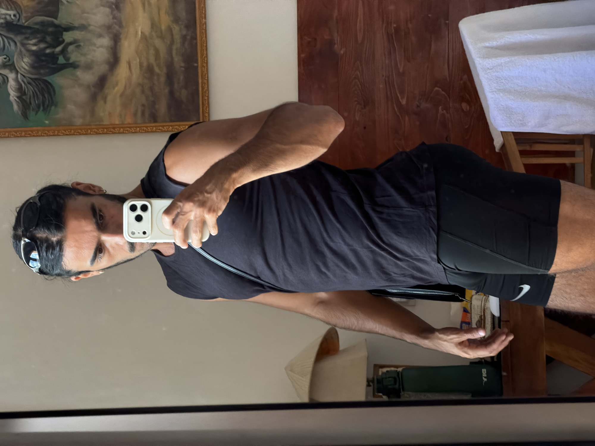 A man is taking a selfie in a mirror. He is wearing a black tank top and black shorts with a white Nike logo. He has dark hair, sunglasses on top of his head, and a beard. He is holding a white iPhone in his right hand. In the background, there is a painting with a gold frame, wooden walls, a white towel on a wooden chair, a green object, and a conical Asian hat. Based on visual information, the location could be North Kuta, Indonesia.