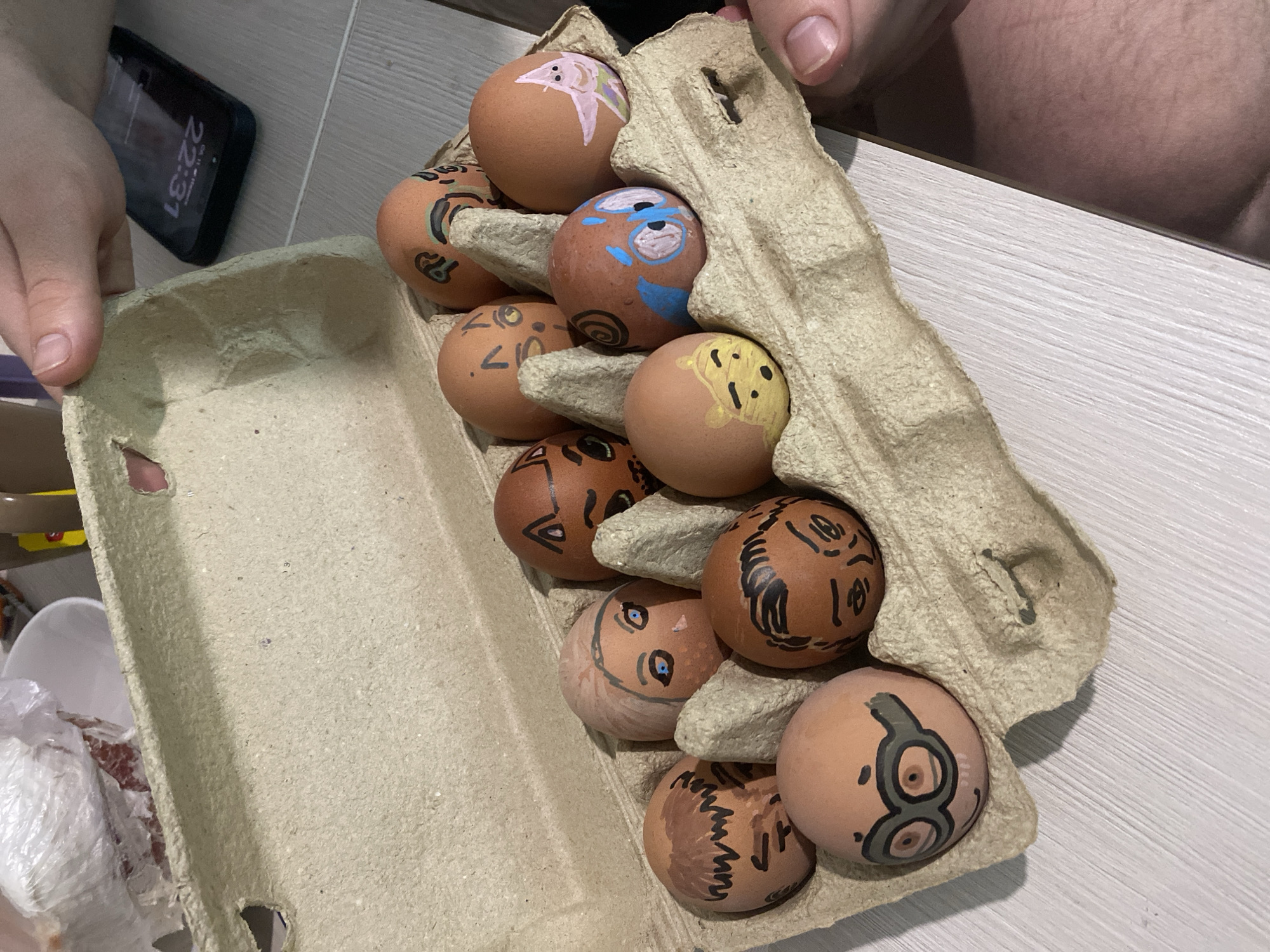 The image shows an open egg carton filled with decorated brown eggs. The eggs are adorned with various cartoonish faces and abstract designs in multiple colors. A hand holds the open carton, which is sitting on a light-colored, possibly wooden, surface. A black smartphone displaying the time "22:31" is visible next to the carton.
There is no indication of Izhevsk, Russia in the images.