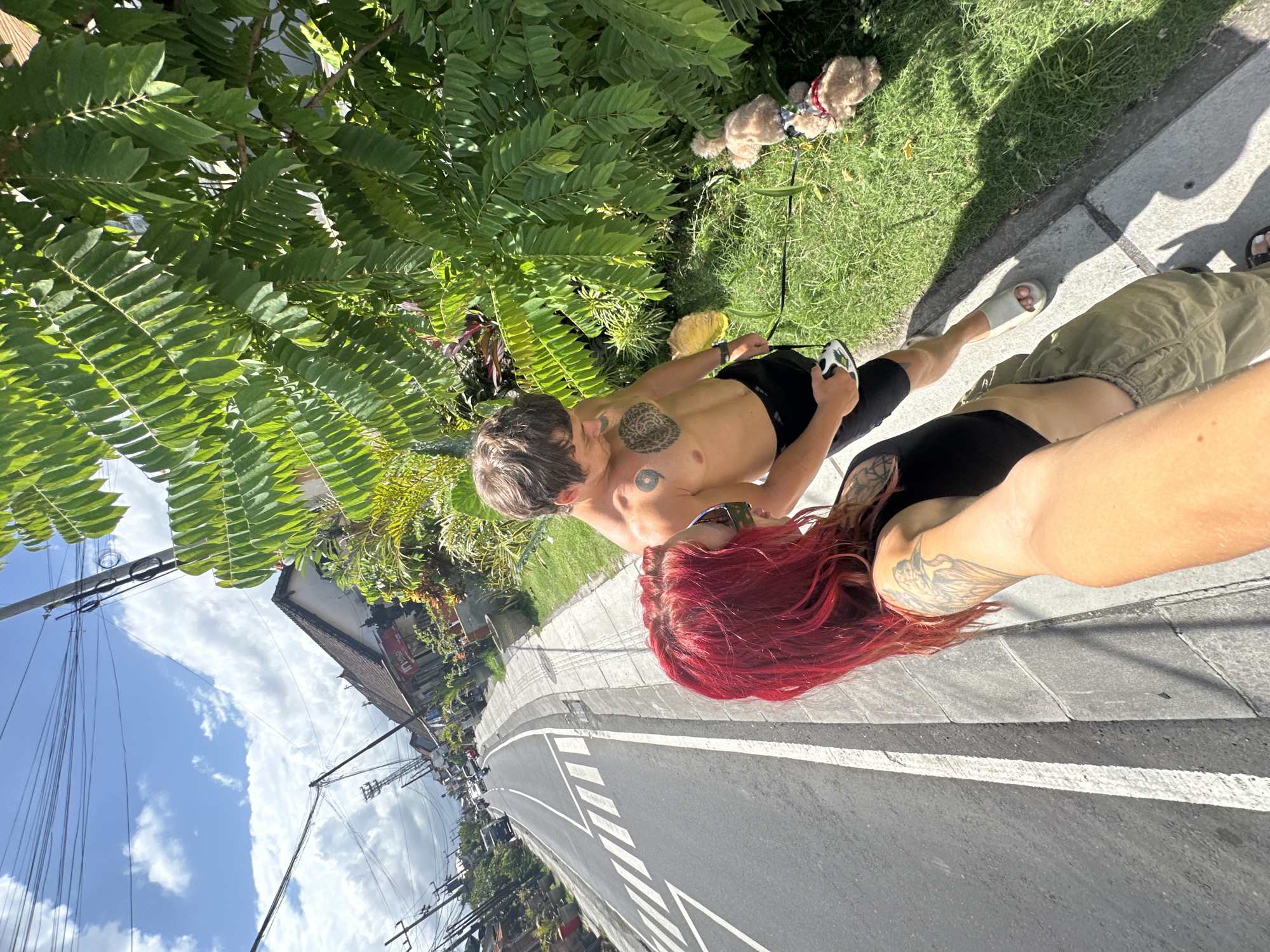 Two individuals, a man and a woman, stand on the edge of a street in North Kuta, Indonesia. The man is shirtless, wearing black shorts and sandals, and has tattoos on his chest and arm. He holds a black leash attached to two stuffed animal dogs placed on the grassy area adjacent to the sidewalk. The woman has bright red hair and wears a black top and olive-colored pants. She also has a tattoo on her arm. They stand on a gray concrete sidewalk bordering a two-lane asphalt road with white lane markings. Lush green vegetation is visible along the roadside, and buildings, power lines, and a blue sky with clouds are in the background.