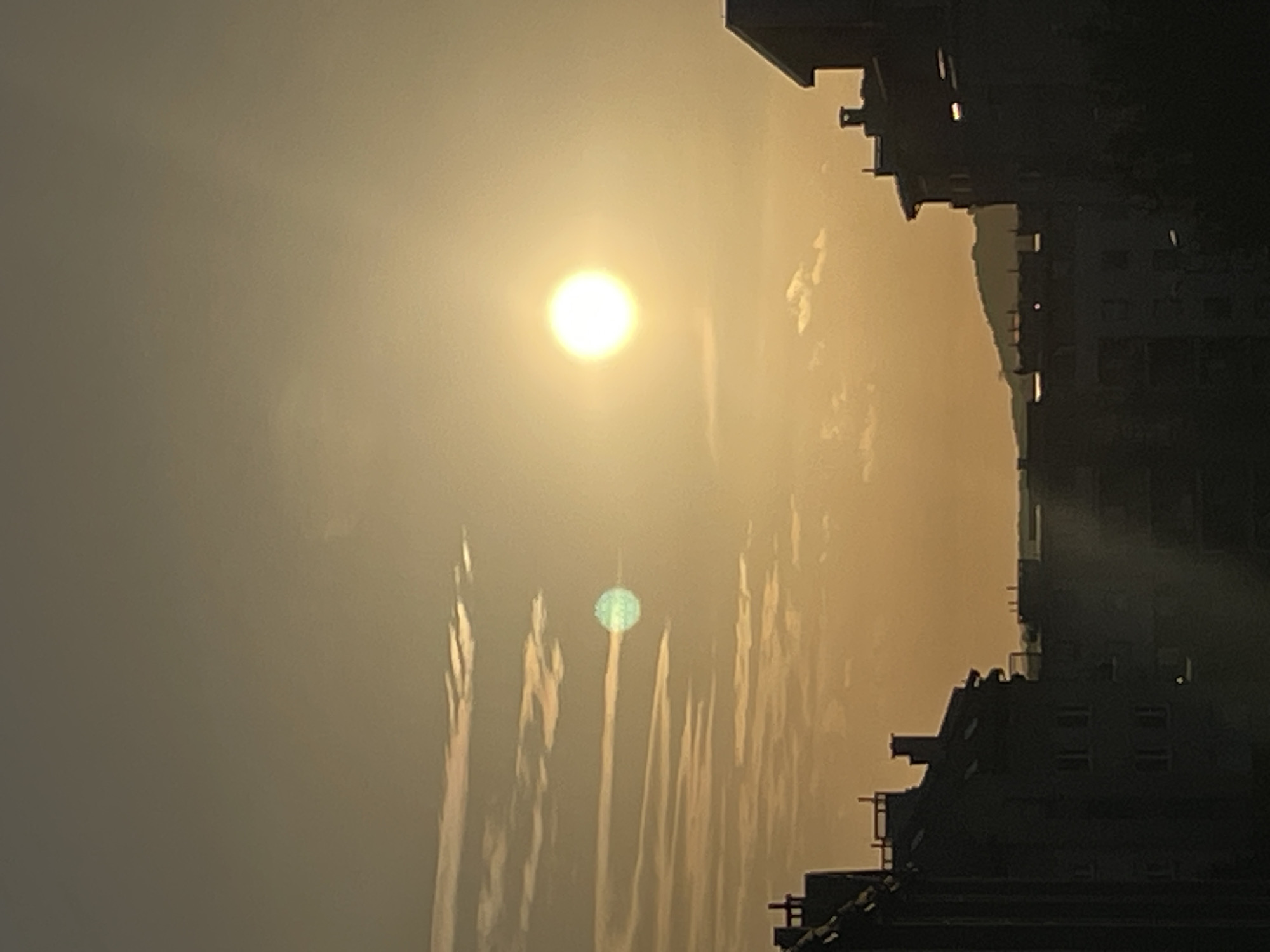 The image depicts a city skyline at sunset. The sky is a warm, hazy orange with wispy clouds. The sun is a bright, orb-like disc in the upper right quadrant of the sky. In the middle left of the sky, there is a greenish, lens-flare-like anomaly. Silhouetted against the sky are the rooftops of several buildings, appearing dark and indistinct. The overall impression is one of a tranquil, urban sunset.