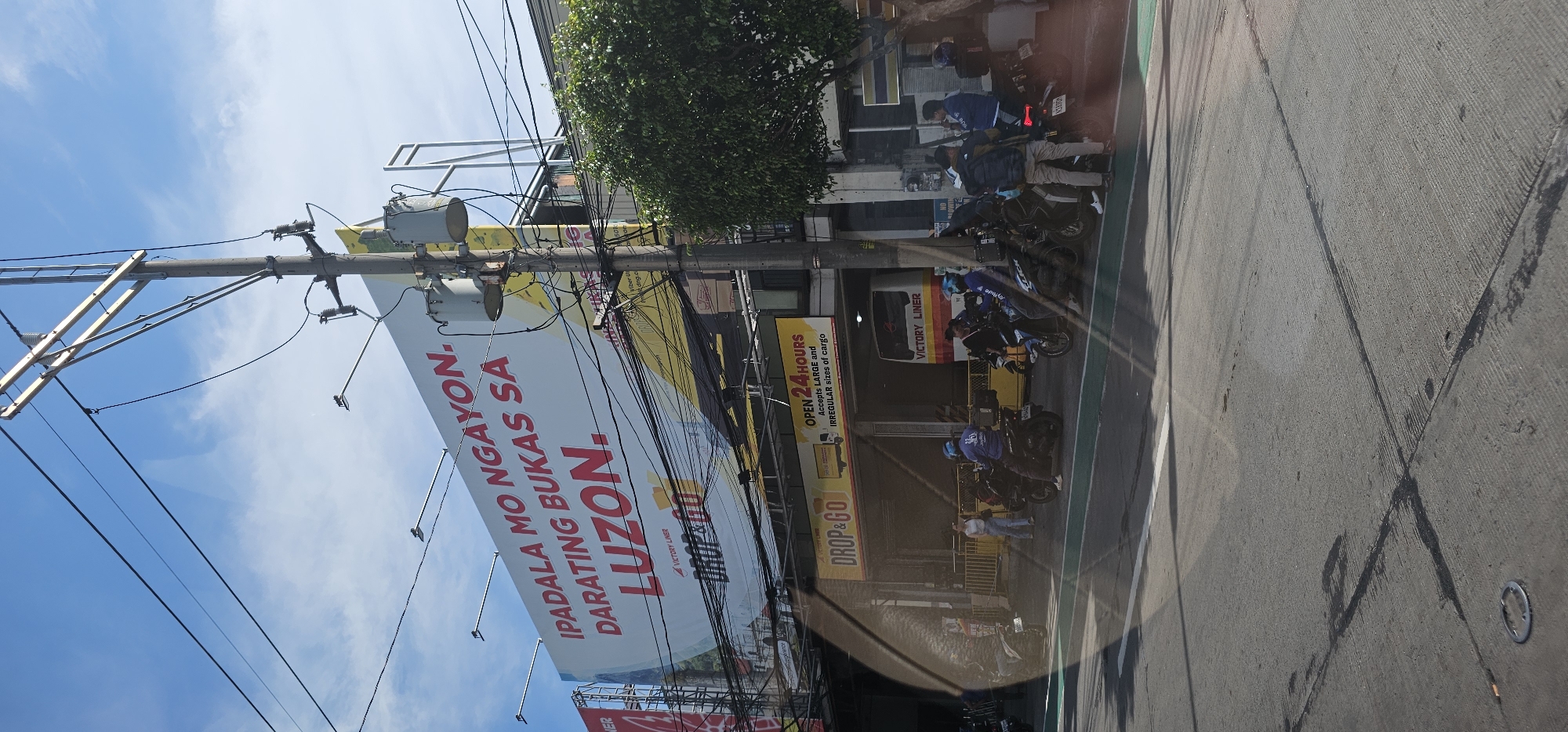 In Quezon City, Philippines, a billboard for Victory Liner's "Drop & Go" service is prominently displayed, advertising "IPADALA MO NGAON, DARATING BUKAS SA LUZON," meaning "Send today, arrive tomorrow in Luzon." Below the billboard, a Victory Liner bus terminal is visible, with several motorcycles parked and people on them, suggesting a waiting area or pick-up point. The scene is characterized by a busy street with utility poles and overhead wires, indicative of an urban setting. The sky is clear with some clouds, and the overall lighting suggests daytime. The presence of a bus and multiple motorcycles implies transportation activity.
