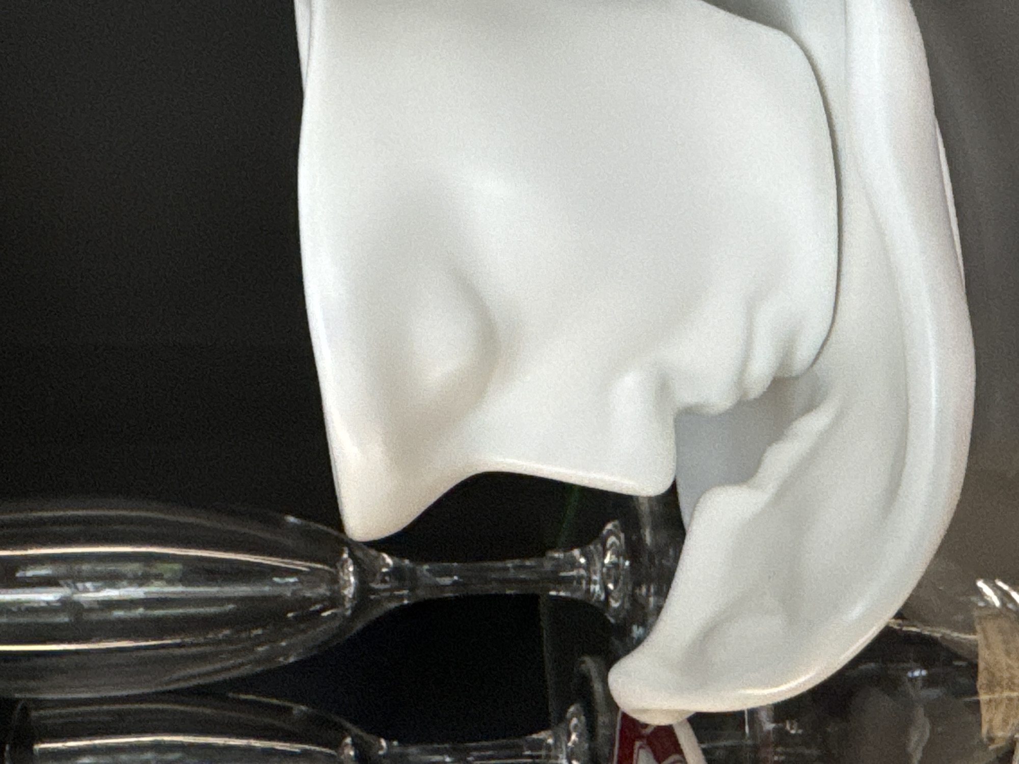 A close-up, side-profile shot showcases a white ceramic mug designed to resemble a human face, complete with sculpted features like a nose, closed eyes, and lips. The mug rests on a matching white saucer, also sculpted to suggest a reclining form. In the background, two clear stemmed glasses, likely champagne flutes, are partially visible. The lighting highlights the glossy texture of the ceramic. The image is an indoor shot, with a dark, out-of-focus background. No specific location within Steklyannyi, Russia, is indicated in the visual data.