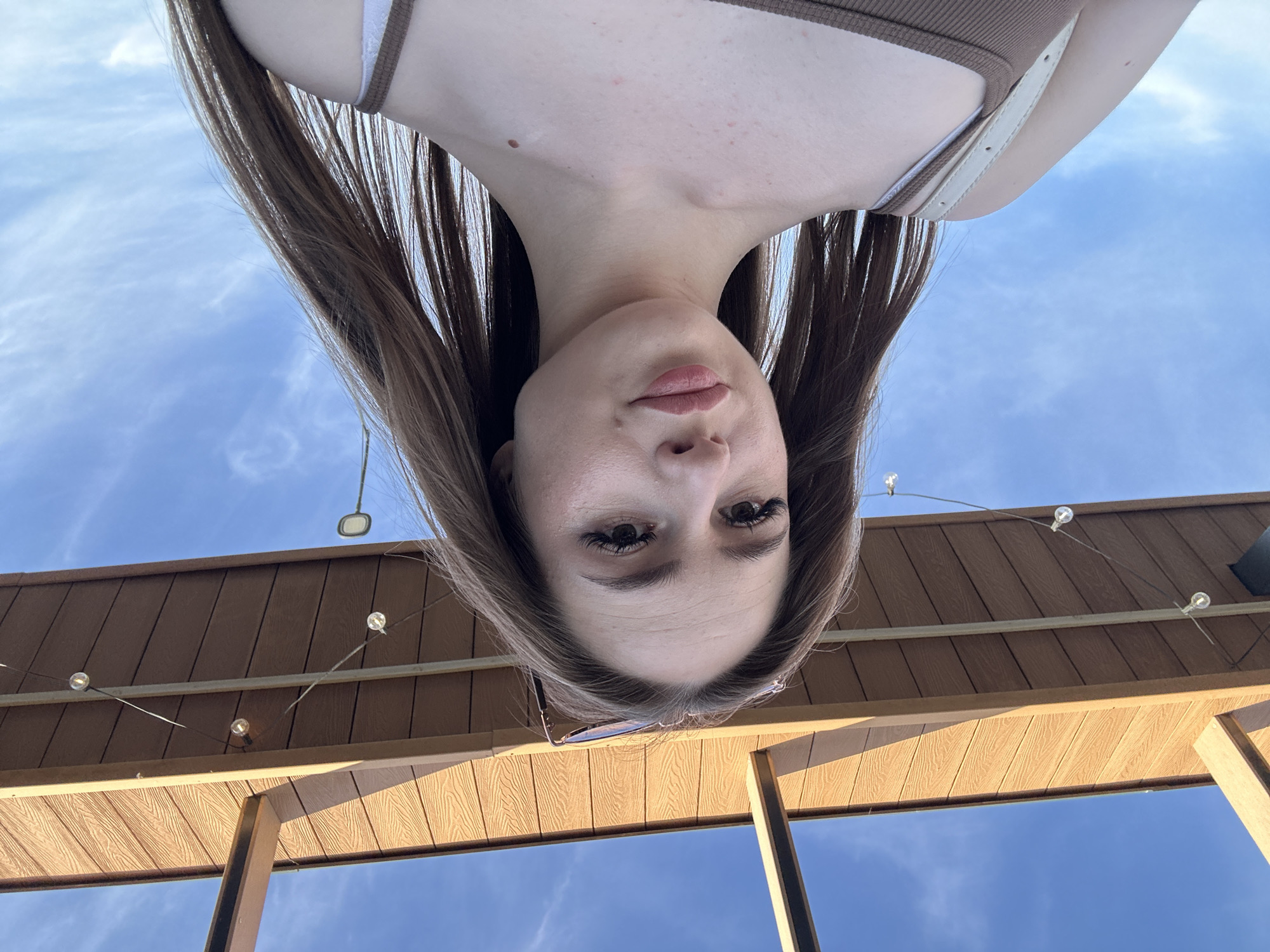 An inverted portrait features a young woman with long, dark brown hair and a neutral expression, gazing towards the camera from an overhead perspective. Her face and upper chest are visible, with her head appearing upside down in the frame. She wears a brown ribbed top, and a white strap is visible on her left shoulder. Her lips are slightly parted, and her eyes, with noticeable eyelashes, are open.
The background primarily consists of a clear, bright blue sky with scattered wispy clouds, suggesting daytime conditions. Below her head, a wooden structure, possibly a pergola or an awning, is visible. This structure is constructed from horizontal planks with a light brown, textured finish. String lights with small, spherical bulbs are strung across the wooden beams. On the right side of the frame, a darker vertical beam of the structure is visible. The bright lighting on the wooden elements indicates a sunny outdoor setting, likely late afternoon or early evening. No discernible text is visible in the image.