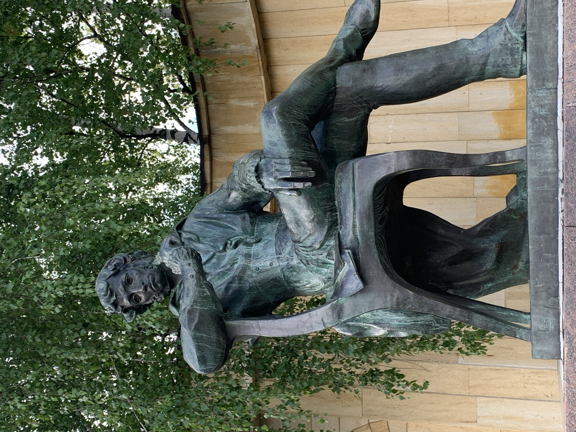A bronze statue of a man, seemingly in deep thought, is reclining in a chair in Stavropol, Russia. His right hand is positioned near his face, and his legs are crossed, with one foot resting on the chair. The statue is situated outdoors, with foliage from a tree partially obscuring the upper left portion of the image. The statue is placed against a building with tan-colored stone exterior. The base of the statue is adorned with dark gray granite.