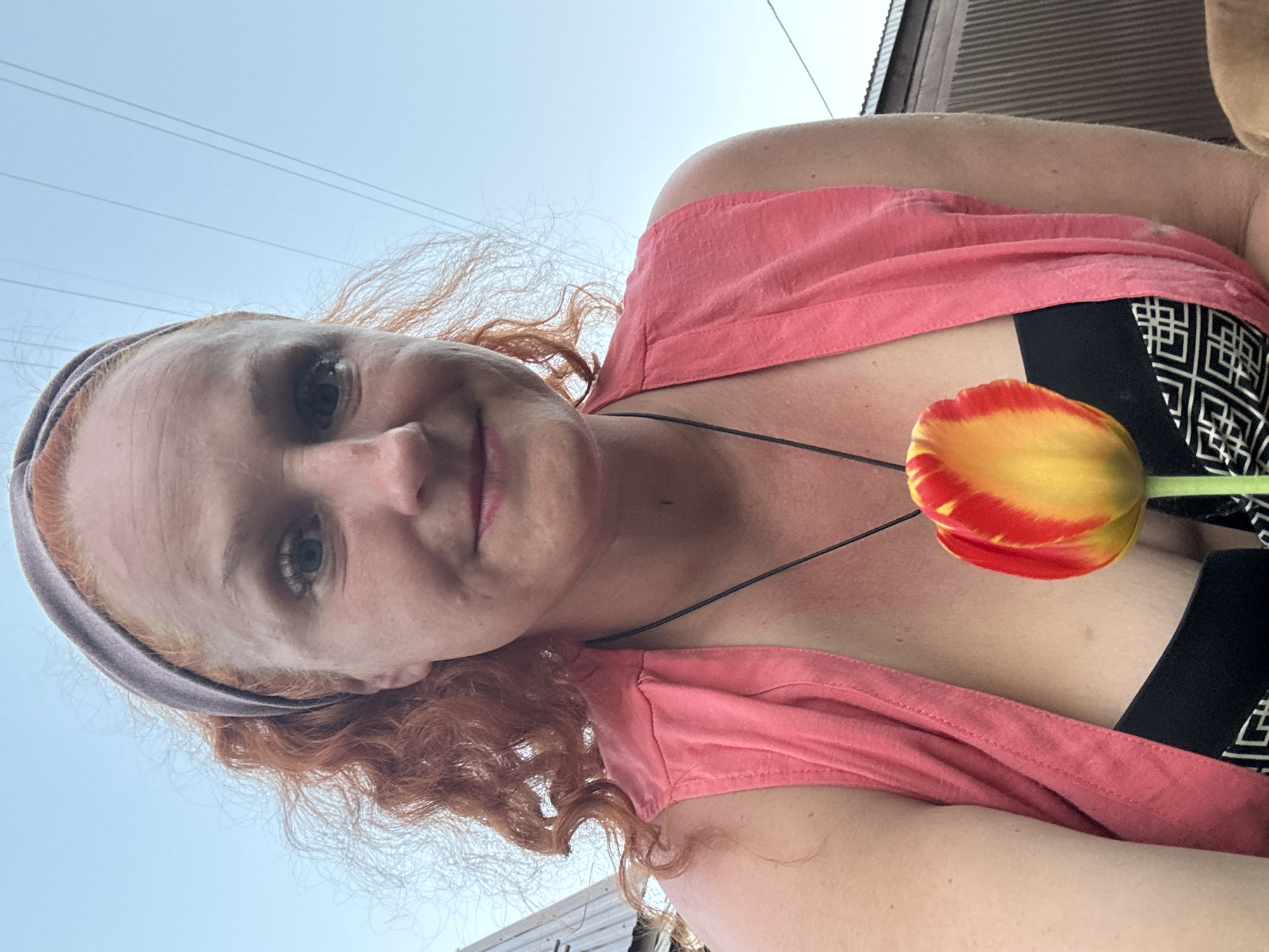 A selfie shows a woman with fair skin and red curly hair, wearing a gray headband. She is smiling slightly, and has on a pink top over a black and white patterned garment. A red and yellow tulip is positioned near her neckline. The background includes a blue sky with power lines. The setting appears to be outdoors, with a partial view of a building. There is no specific information indicating the location is in Chaikovsky, Russia.