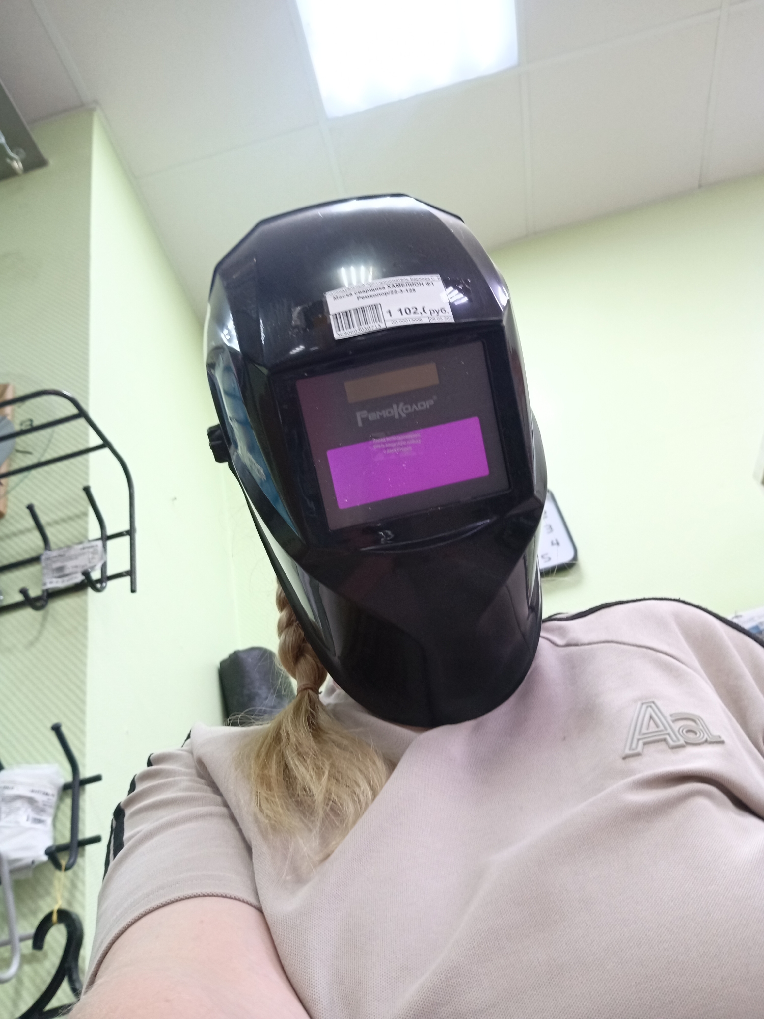 A person is wearing a black welding mask in what appears to be a retail environment. The mask has a price sticker that says "1 102 руб" and the text "РемоКолор" on the viewing window. A white or light-colored sweatshirt with the letters "Aa" is visible below the mask. The person has blonde hair pulled back in a ponytail. The background shows a light green wall, a white tiled ceiling with fluorescent lighting, and retail shelving to the left. The location appears to be in Фоки, Russia, based on the Cyrillic text on the mask and the price sticker.