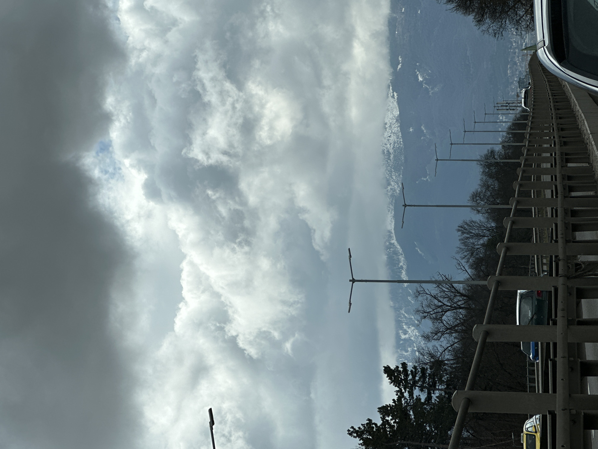 The image presents a view from a bridge or elevated position in what appears to be the city of Sofia, Bulgaria. The foreground shows a metal railing and partially visible cars. The mid-ground features a row of streetlights extending into the distance, interspersed with bare trees. Beyond the trees, snow-capped mountain peaks are visible against a predominantly cloudy sky. The clouds range from dark and ominous to brighter, lighter formations.