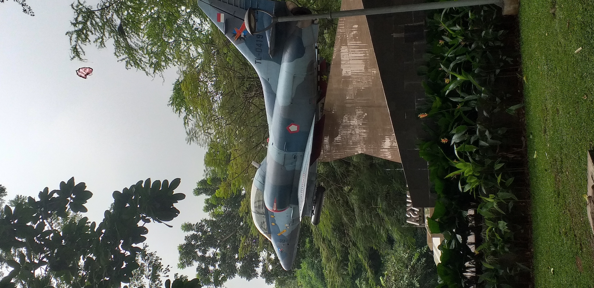 The image depicts an A-4 Skyhawk aircraft mounted as a monument in a park setting. The aircraft is painted in camouflage colors, with Indonesian flags visible on its tail. The plane is positioned on an angled steel pylon. Lush green trees and foliage surround the monument. A small, faded object is visible in the upper-left sky area. The monument is likely located in the city of South Jakarta, Indonesia, based on the A-4 Skyhawk monument in Menteng Park.