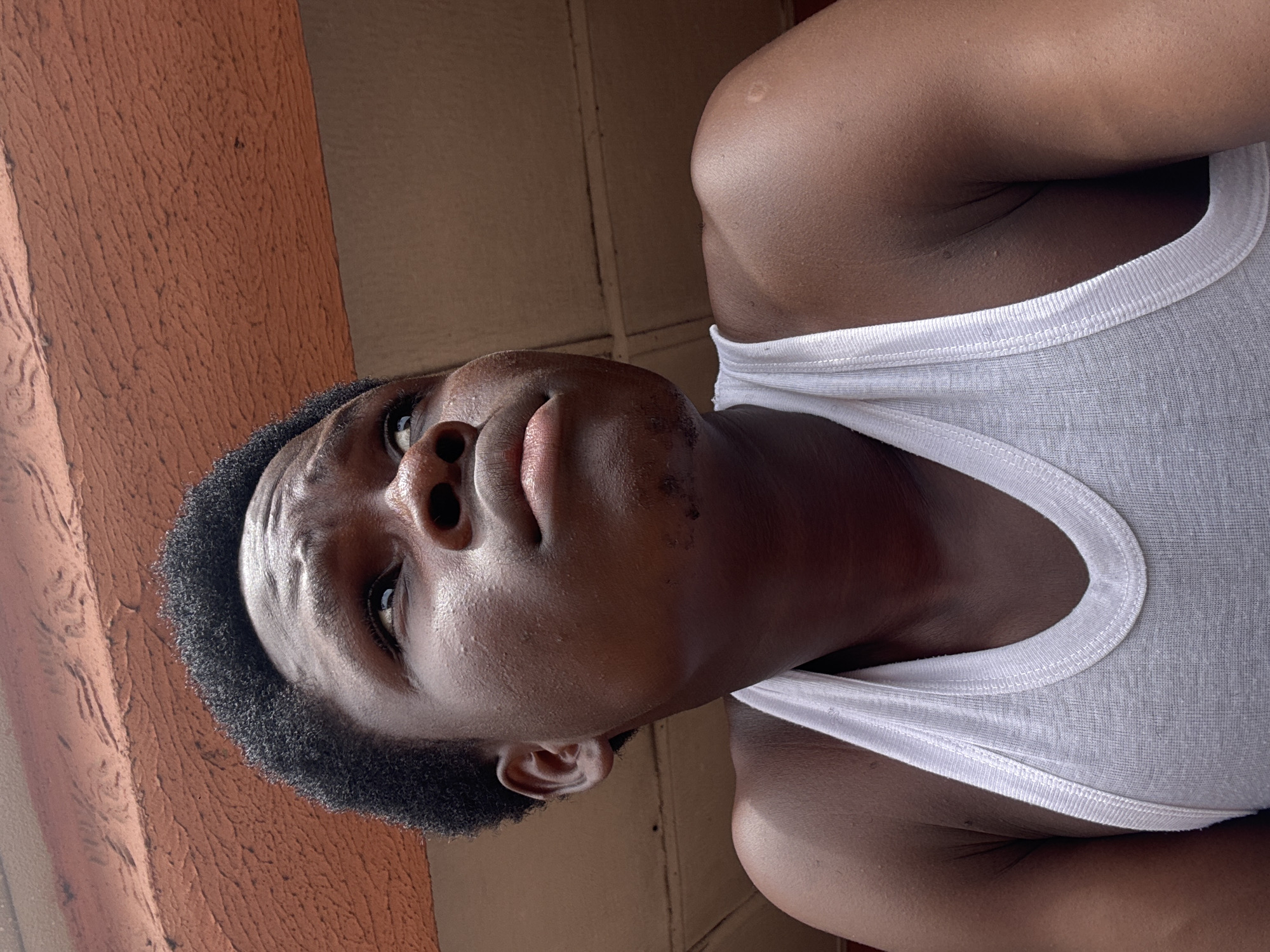 A low-angle portrait of a Black man wearing a white tank top. He is looking upwards. The background consists of a brown wall on the right and an orange wall on the left. The location cannot be determined from the image.
