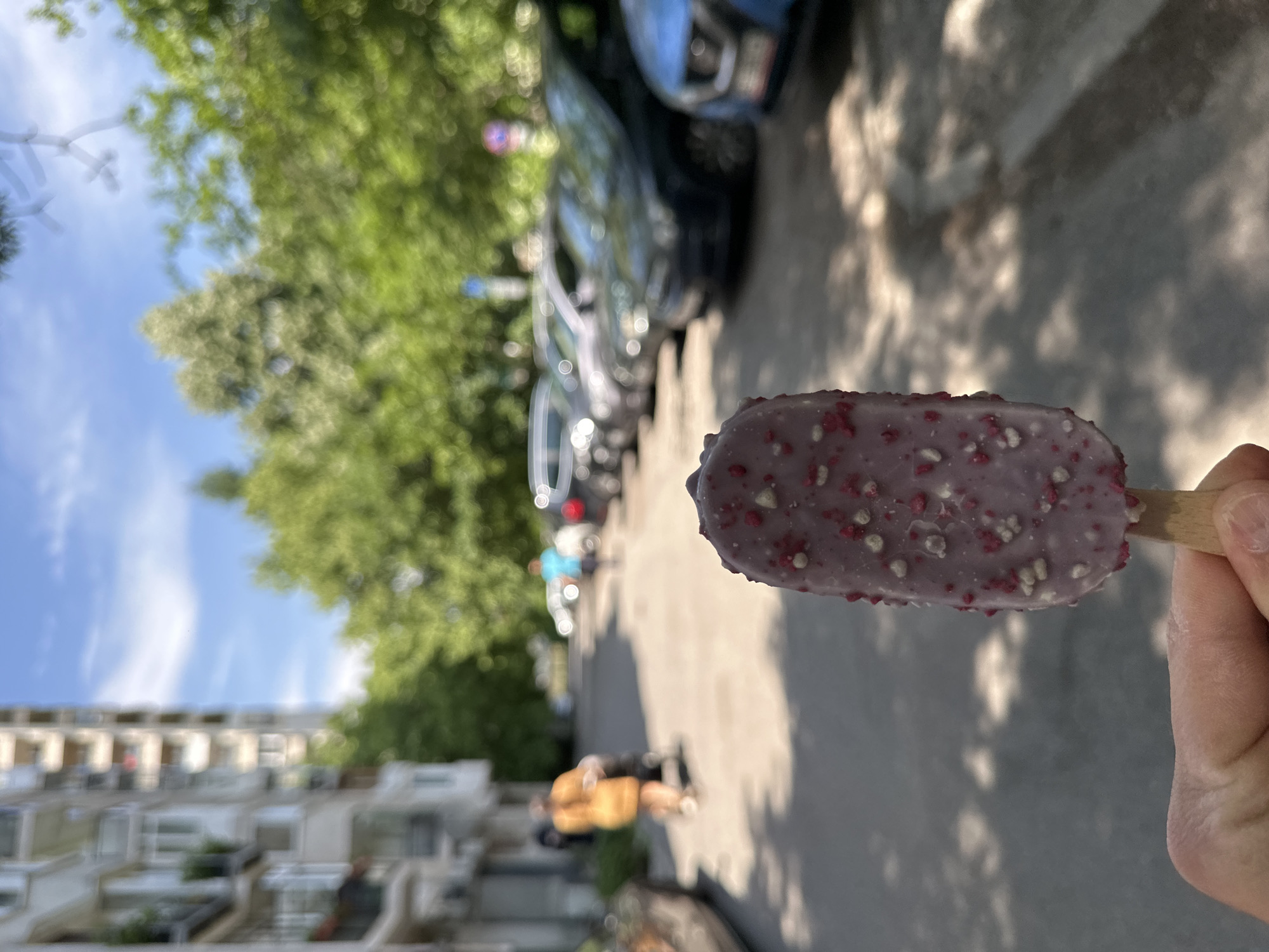 A close-up, slightly tilted, eye-level shot focuses on a rectangular purple popsicle held by a hand, seemingly in an outdoor urban setting. The popsicle features small white and red sprinkles on its surface.
In the background, out-of-focus elements create depth. A row of parked cars lines the right side of the frame, casting dappled shadows on the gray asphalt road. On the left, trees with green foliage fill the frame, partially obscuring white residential buildings under a bright sky.
Further back, indistinct figures of people can be seen walking on the sidewalk. The location appears to be a street scene in Sofia, Bulgaria, identified by the architectural style of the buildings and street layout. The overall impression is a sunny day in a residential area, with the primary subject being the popsicle.