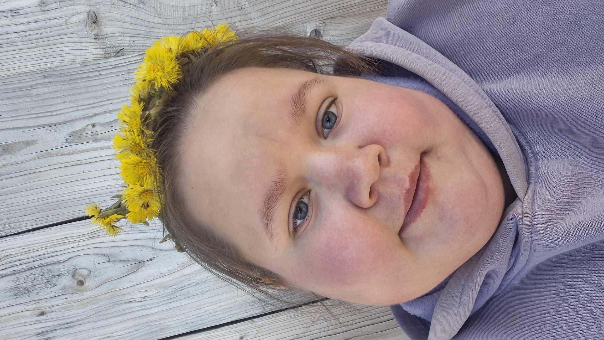 A medium, angled, close-up shot shows a light-skinned female with blue eyes and a flower crown, positioned on a light-colored wooden surface. She has a slight smile. She is wearing a lavender hooded sweatshirt. The image provides no discernible information about the location in Дойная, Russia.