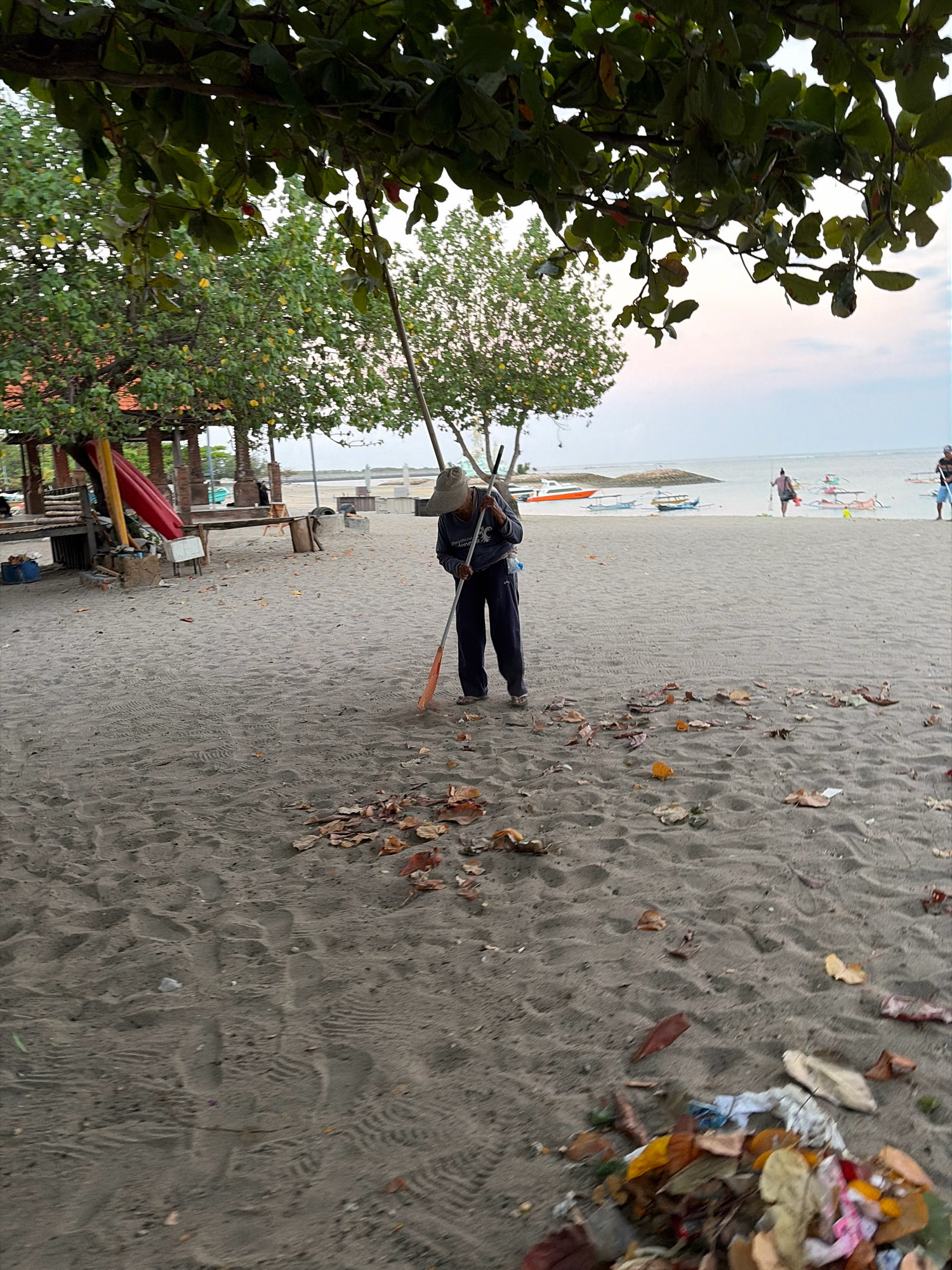 An individual wearing a blue shirt and a light-colored hat is sweeping the sandy beach with a broom. The person is positioned in the mid-ground of the image, facing away from the camera and towards the ocean. Fallen leaves and some debris are scattered on the sand around the individual. In the background, the ocean is visible with several boats docked near the shore. The sky is overcast, suggesting it might be early morning or late afternoon. The scene appears to be a public beach in Kuta, Indonesia, indicated by the presence of typical beach structures and the style of the boats. The overall atmosphere is one of a routine clean-up operation on a tropical beach.