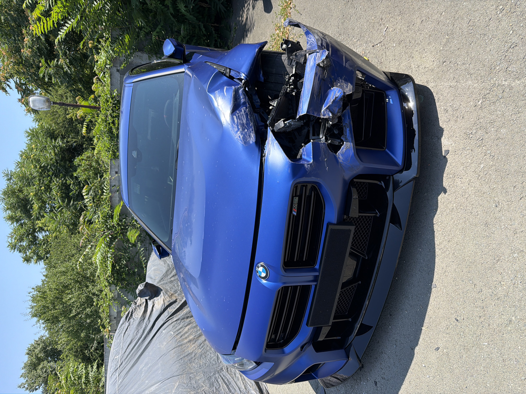 A damaged blue BMW M2 is parked outdoors on a paved surface, likely in Burgas, Bulgaria, judging by the foliage and the style of the lamppost. The car's front right fender and bumper are severely crushed, with parts of the headlight assembly exposed and broken. The hood is slightly ajar on the right side. The vehicle is a sporty coupe with prominent front grilles and a black splitter. In the background, a concrete wall is visible, topped with lush green trees and bushes. A large, dark gray tarp covers another object to the left of the car. A tall, slender lamppost stands among the trees behind the car. The sky is clear and blue. The scene appears to be a street or an open lot in a sunny, urban environment.