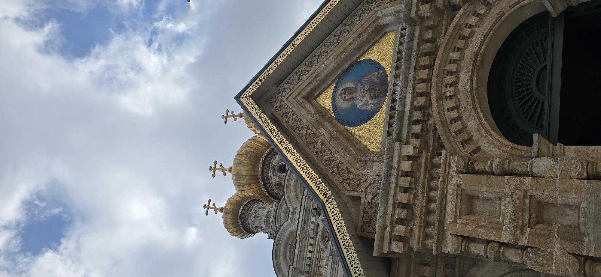 The image shows the exterior of a building with golden domes and intricate stone carvings. The building appears to be a church, with crosses atop the domes. In the center of the facade, there is an oval mosaic of a woman in white robes with a halo. The building is made of light-colored stone. The sky above is cloudy. This is a view of the Church of Mary Magdalene in Jerusalem, Israel, a Russian Orthodox church located on the Mount of Olives.