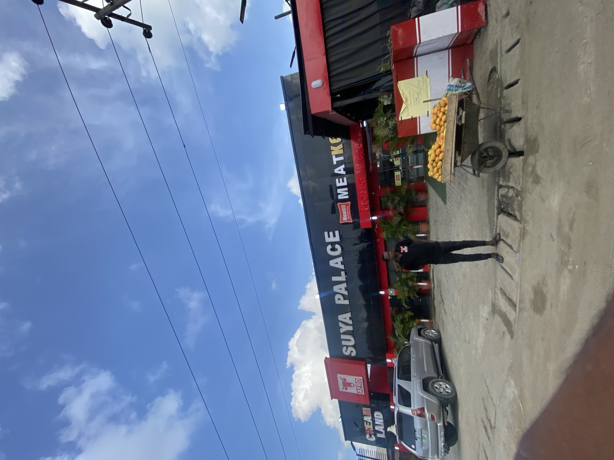 The image shows the "SUYA PALACE" restaurant in Ado, Nigeria, as indicated by "CREAM LAND" and "AFRO" on the building. A man wearing a black shirt and pants stands on the paved area in front of the restaurant. To his right is a cart loaded with mangoes. A silver SUV is parked near the restaurant. Overhead are power lines against a blue sky with scattered clouds.