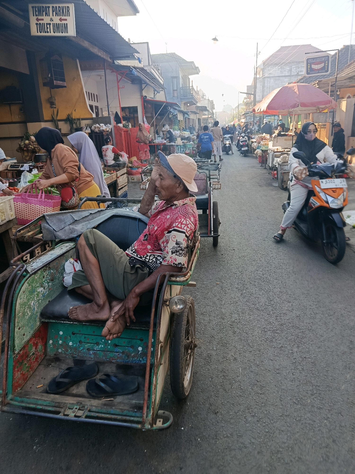 A street scene in Gondanglegi, Indonesia, depicts a busy market with stalls lining the road. In the foreground, an elderly man wearing a red and white patterned shirt, green pants, and a light-colored cap sits in a three-wheeled vehicle, possibly a becak, with his legs exposed. He appears to be resting his head on his hand. Next to him, inside the vehicle, are a pair of black flip-flops and a cigarette butt. The vehicle itself shows signs of wear and tear with peeling green and red paint. Behind him, several women are visible at market stalls. One woman in a brown top and headscarf is arranging items in baskets. Another woman in a light blue headscarf stands nearby. Further down the street, a woman in a white and black patterned top and black hijab rides an orange motorcycle, looking towards the viewer. The street is paved with asphalt and lined with buildings. A red umbrella is open over one of the market stalls. Signs are visible on some buildings, including one in Indonesian that reads "TEMPAT PARKIR & TOILET UMUM." The overall atmosphere suggests a bustling local market in the morning or late afternoon, given the lighting.
