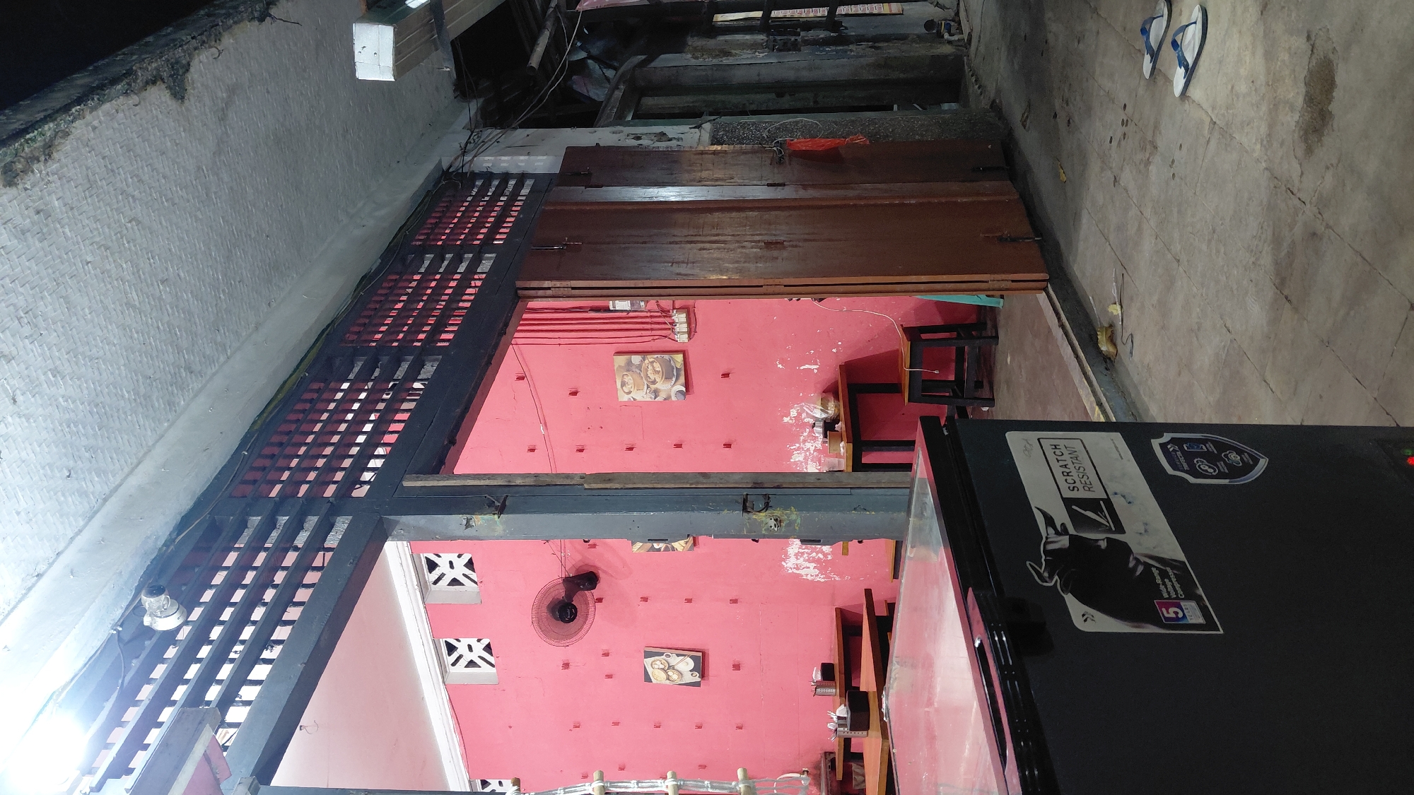 The image depicts the exterior of a food establishment in Bantarsoka, Indonesia, likely a warung or small restaurant. In the foreground, a large, black chest freezer with stickers is visible. The freezer has a "Scratch Resistant" label featuring a bull silhouette. Beyond the freezer, an open doorway leads into the establishment. Inside, the walls are painted a bright pink. Several wooden tables and chairs are set up, with some condiments on the tables. A wall-mounted fan is present on the pink wall, along with framed pictures of food. The establishment appears to be open to the street, with a slatted wooden overhang extending over the entrance area. The ground outside is paved with concrete tiles, and a pair of white flip-flops rests on the ground to the right. The overall environment suggests a casual, possibly bustling, dining setting typical of Indonesia.